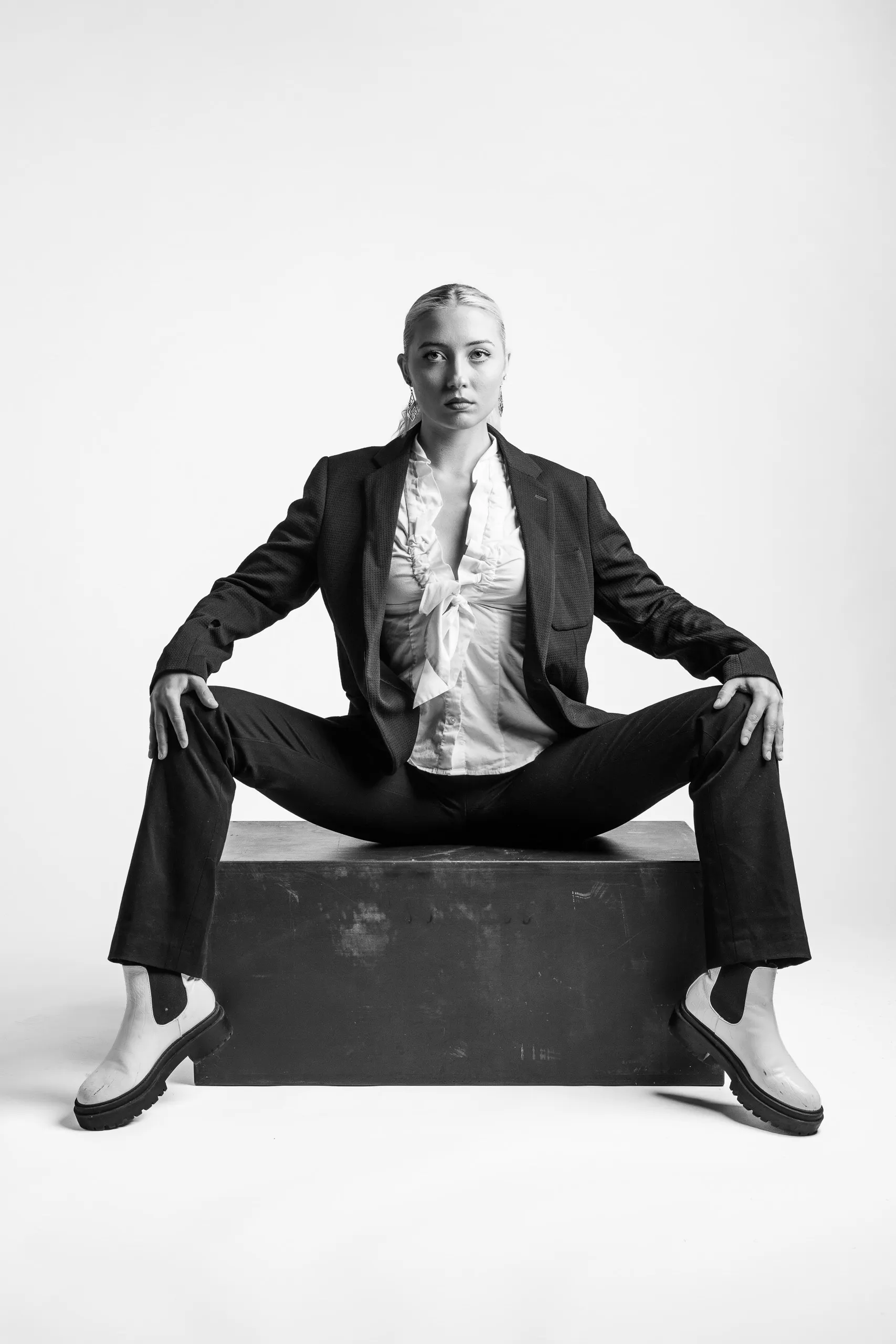 A person with hair pulled back wearing a suit and white shoes sitting on a black box. The person is looking straight ahead.