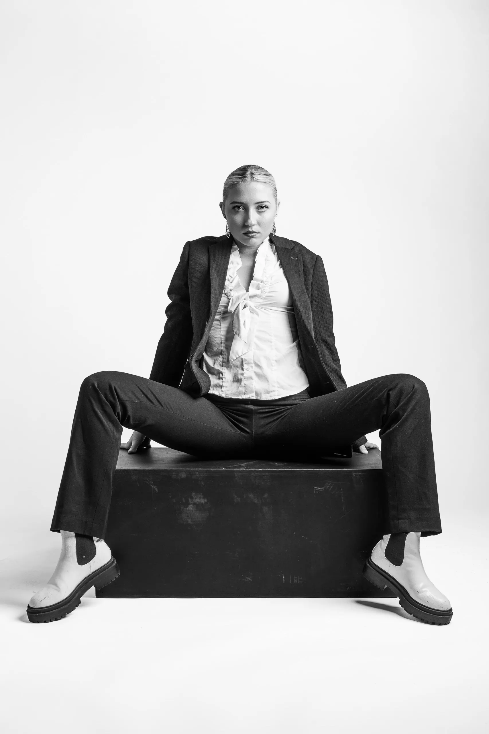 A woman with short hair sits on a black box, wearing a white shirt, a black jacket, black pants, and white shoes. Her legs are far apart as she looks at the camera.