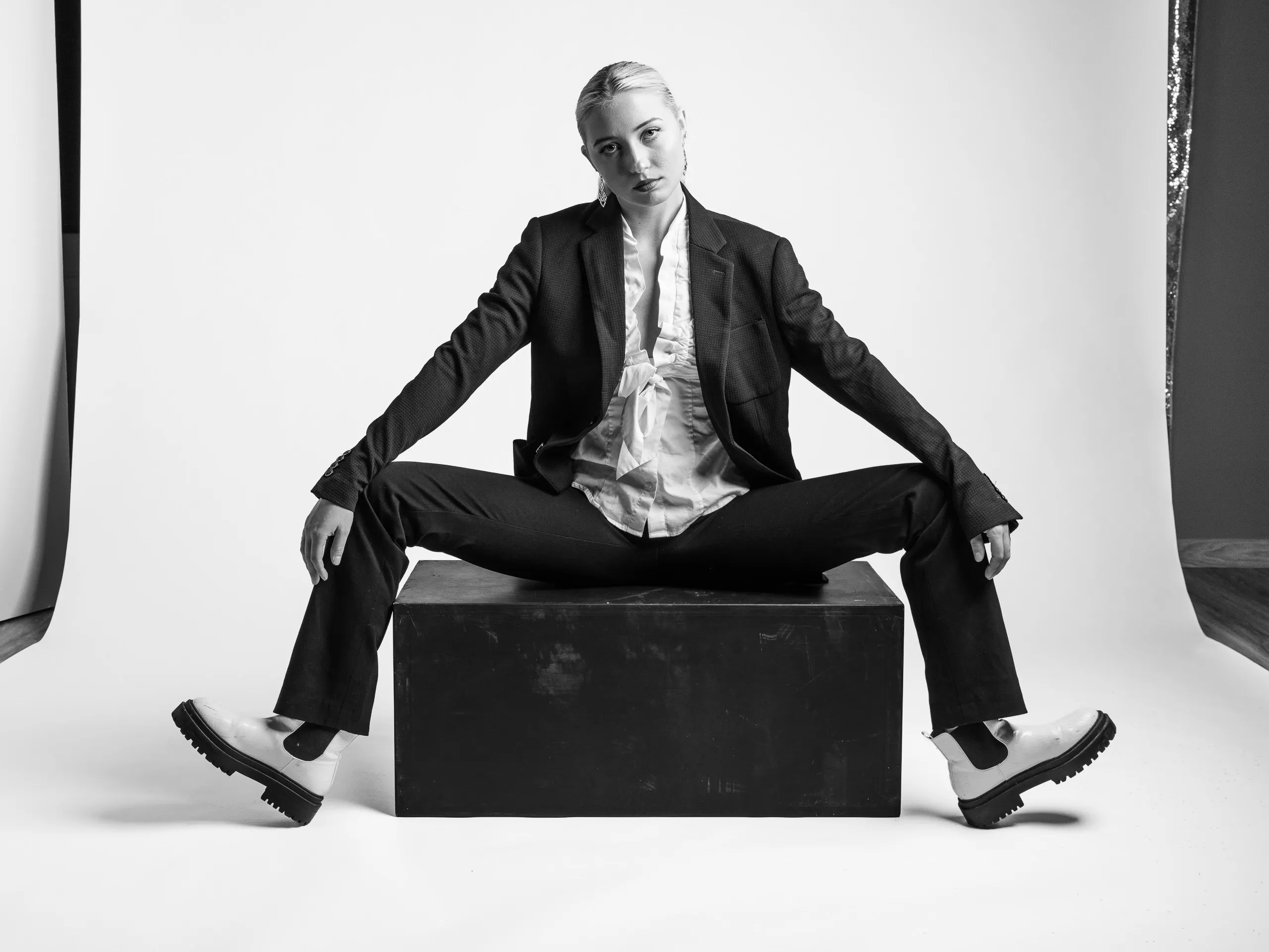 A person wearing a suit and white shoes sitting on a black box. The person is sitting with legs apart and arms resting on the knees.