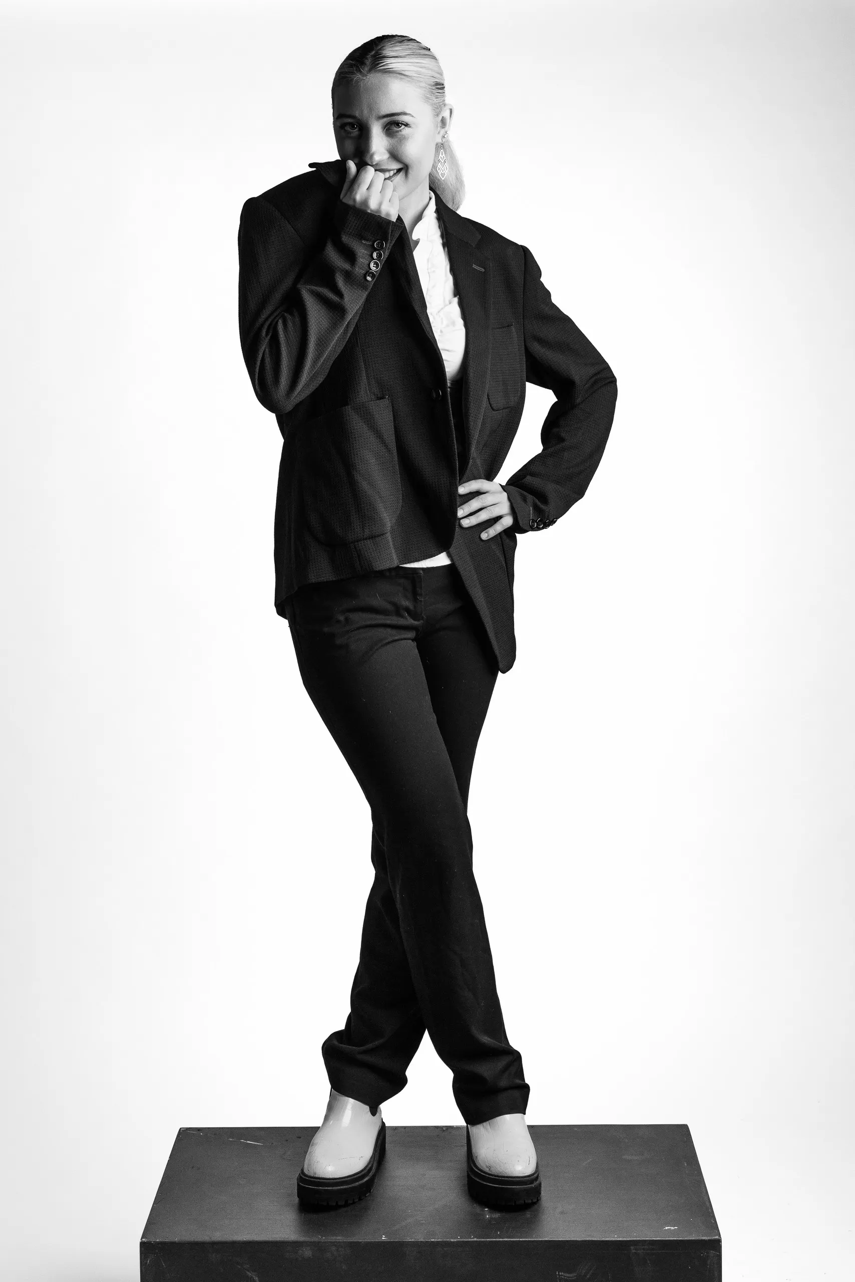 A woman wearing a black suit and white shoes standing on a black block. She is touching her mouth with her hand and standing with one hand on her hip.