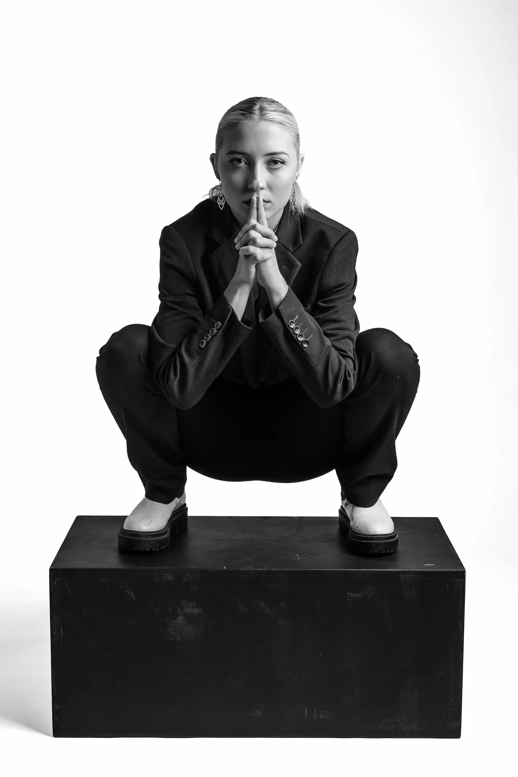 A woman in a suit crouches on a black box, holding a finger to her lips. She has short hair and is wearing white shoes.