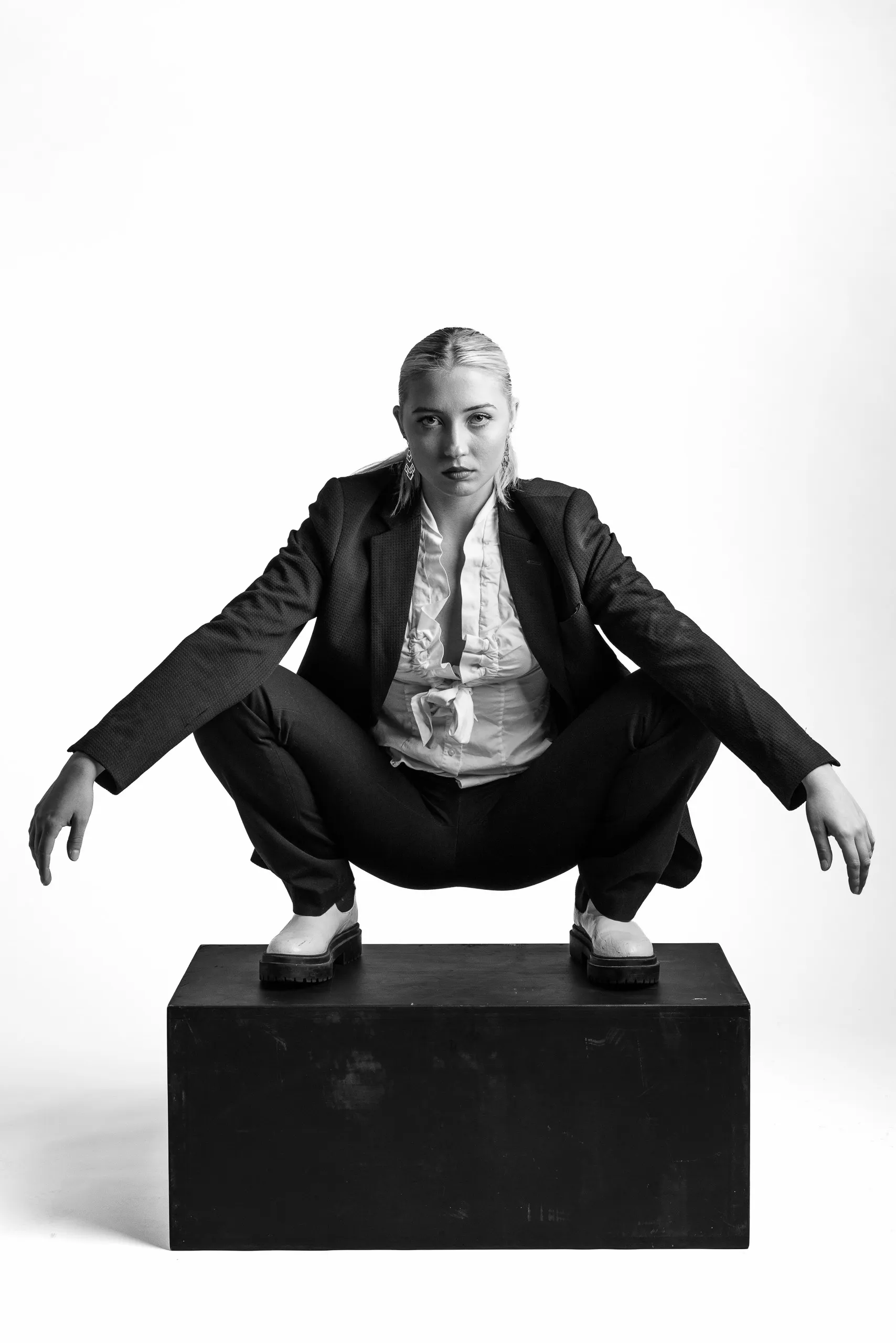 A woman with her hair pulled back squats on a black box. She wears a suit jacket, a patterned shirt, and shoes.