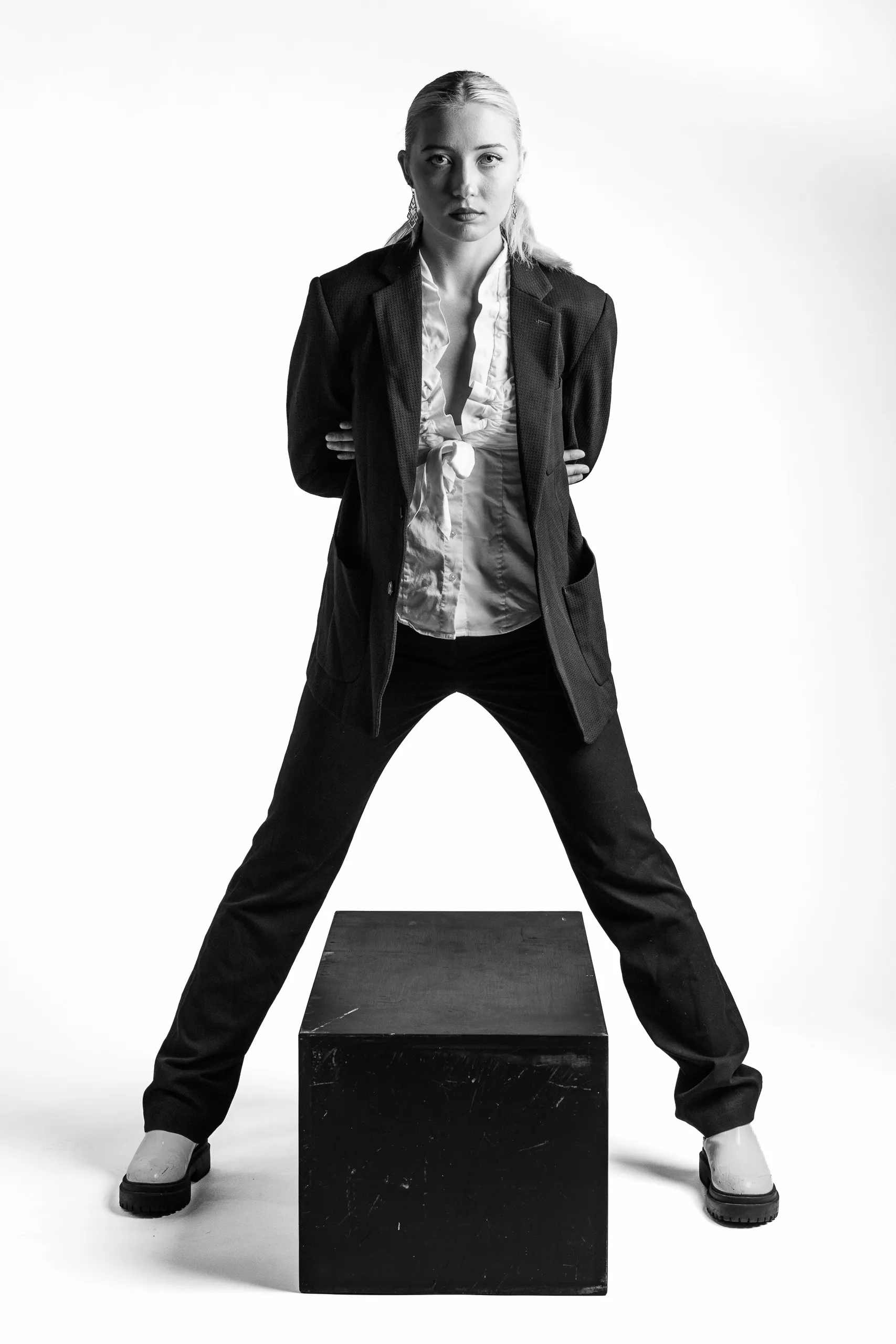 A woman standing with her hands behind her back, wearing a suit coat and black pants, with her legs spread wide apart, standing over a black box.