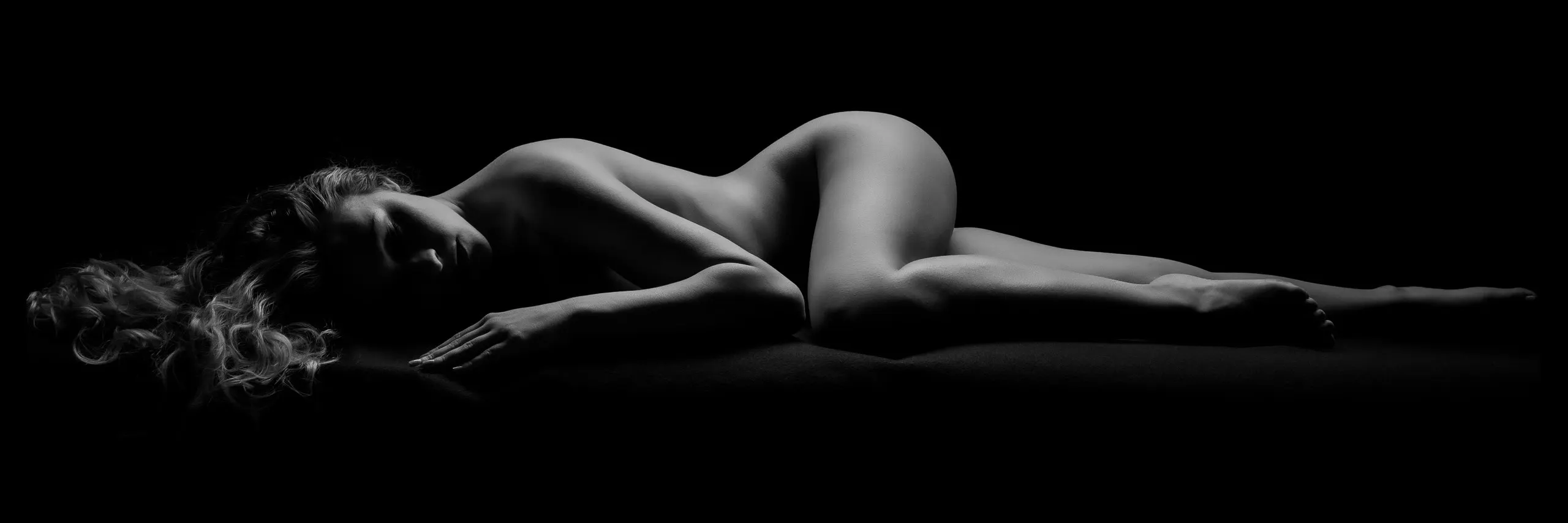 A woman lies curled on her side, hair spilling across the floor in front of her, softly lit in black and white against total black.