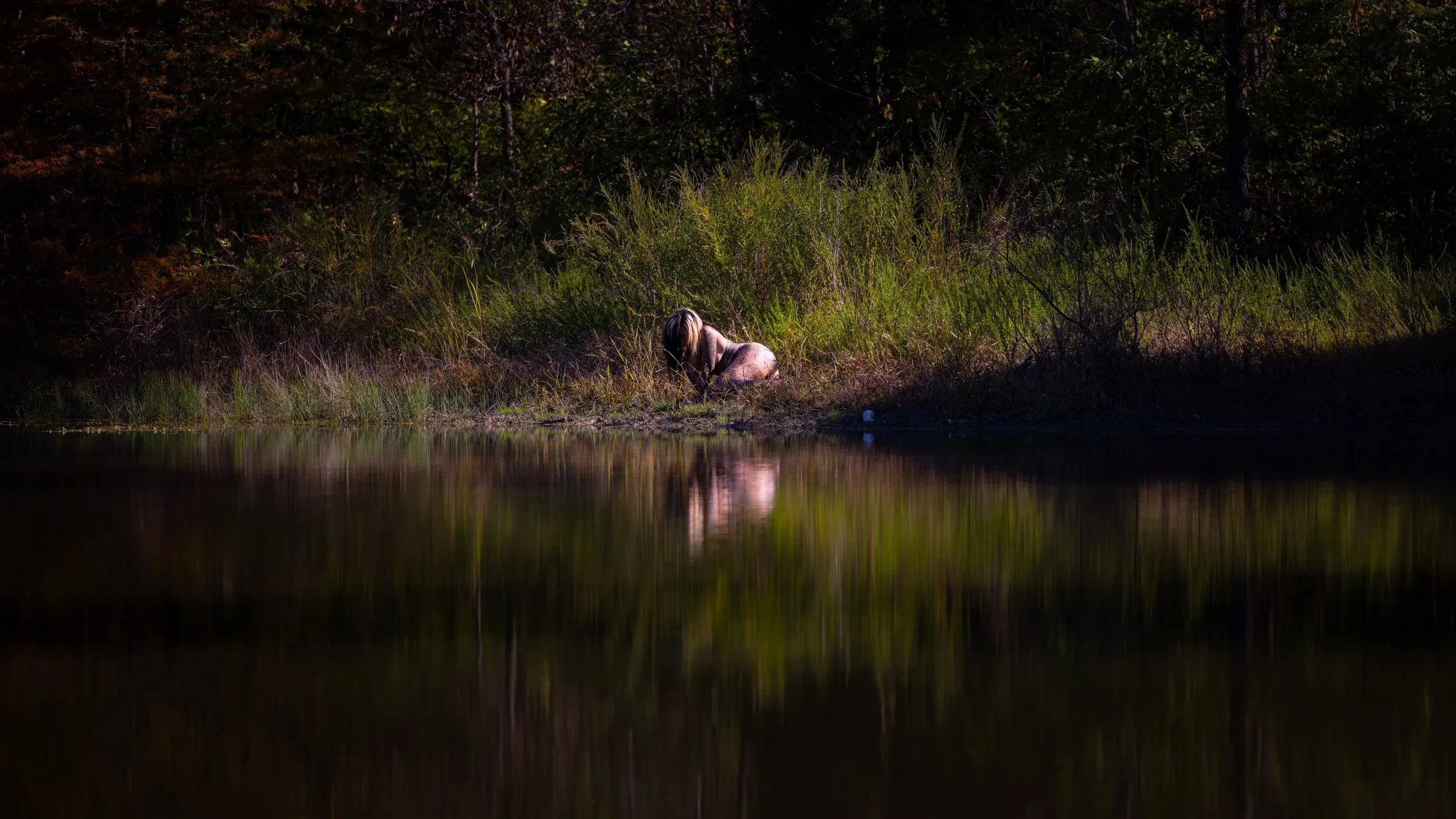 A distant figure crouches beside still water reflecting autumn trees.