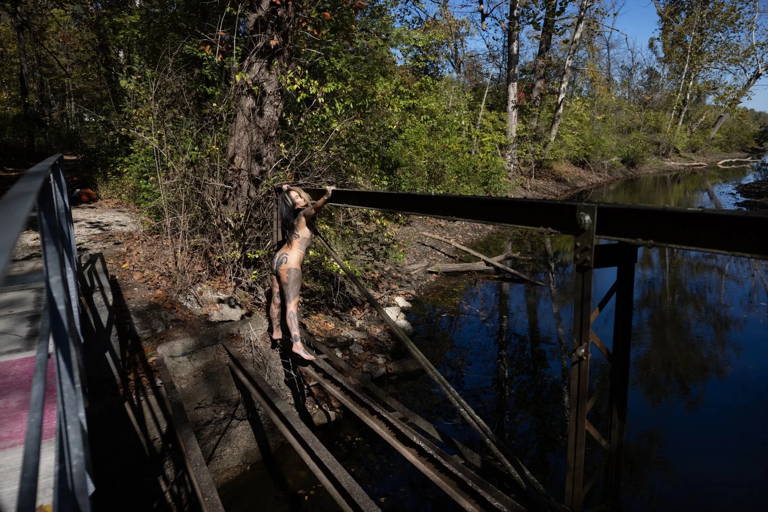 A woman stands amid the beams of a decaying structure beside water.