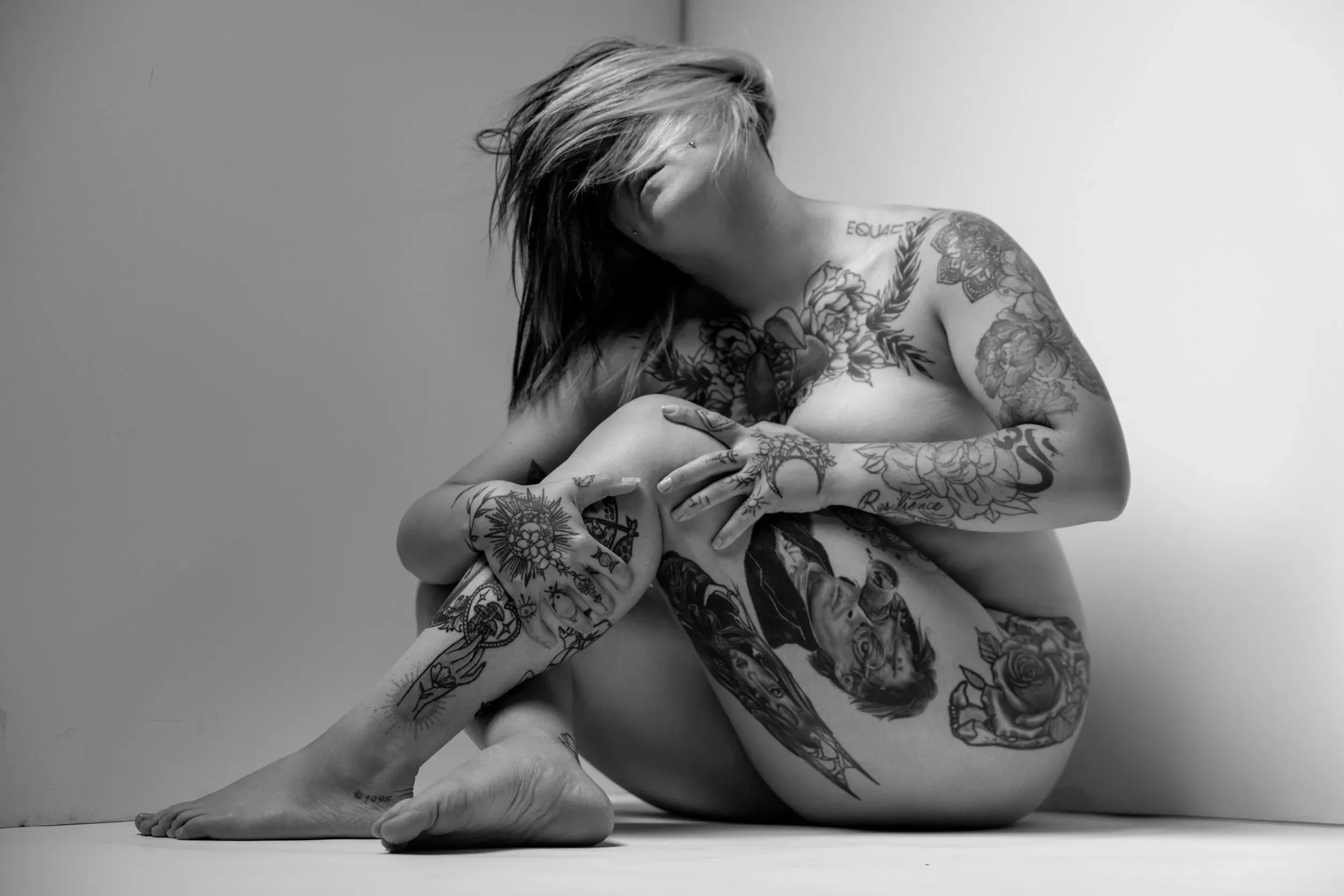 A tattooed woman sits cross-legged against a bright seamless backdrop.