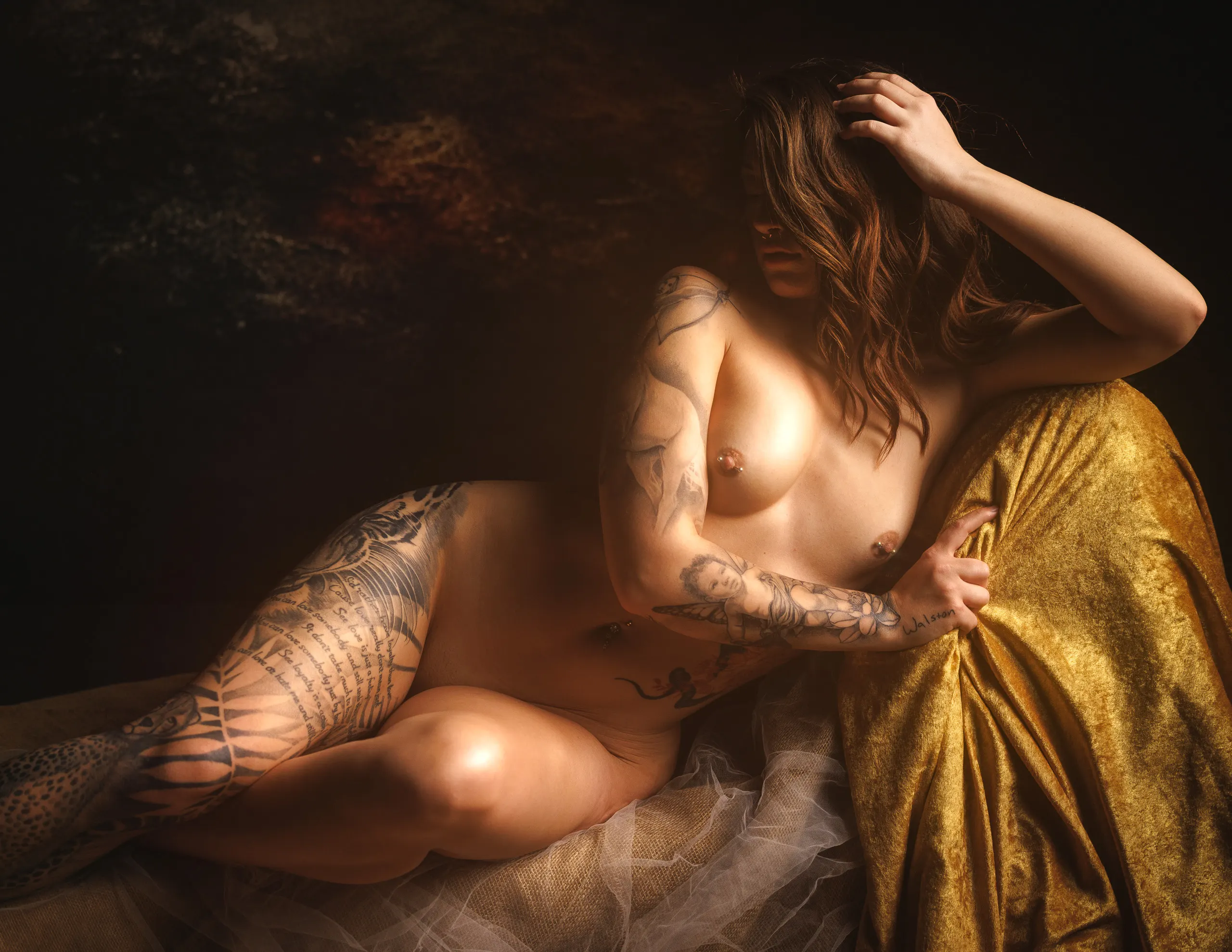 A tattooed woman reclines against gold velvet, one arm raised to her hair, the other cradling her chest, warm firelit light picking out detailed tattoo work along her arm and leg.
