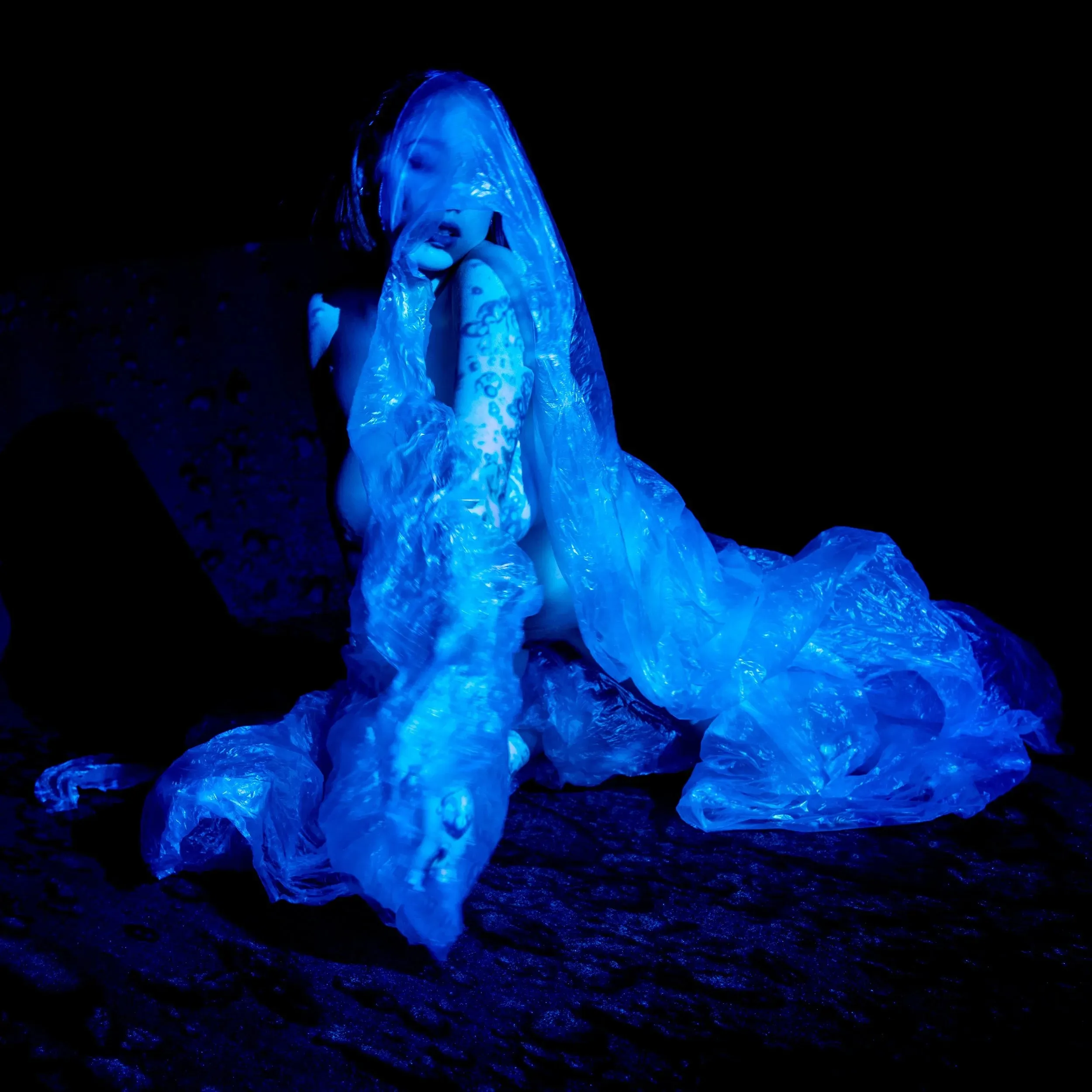 A seated figure wrapped in crinkled plastic glows electric blue against black.