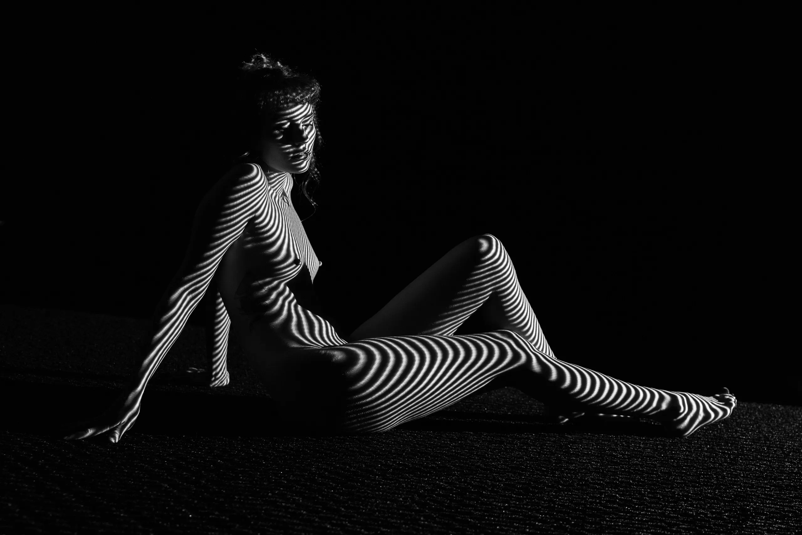 A woman sits with knees drawn to one side, leaning back on one arm, sharply banded stripes of light and shadow wrapping her entire body and legs.