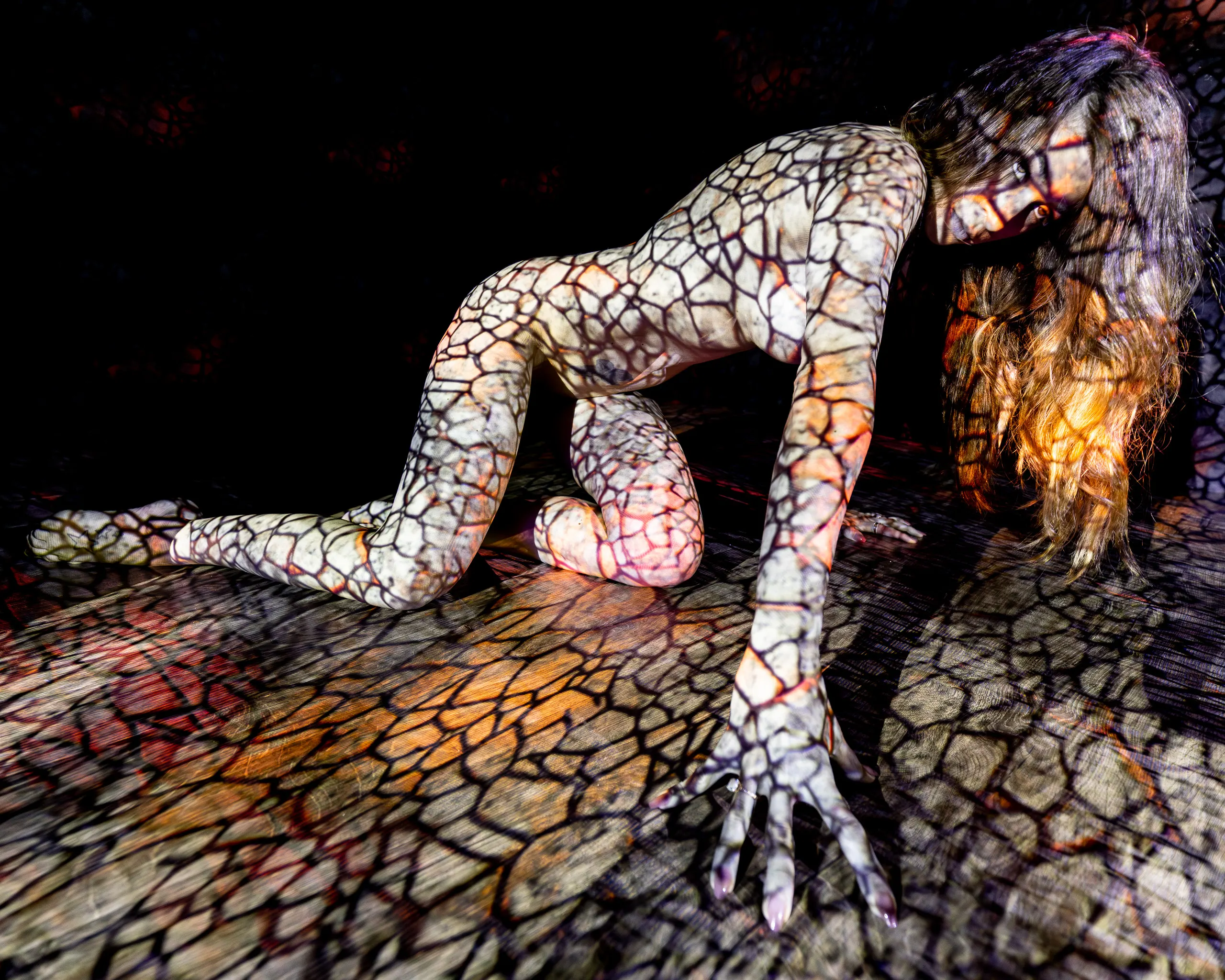 A woman kneels on all fours, a cracked, mosaic-like pattern of light in warm gold and black projected across her whole body and the reflective floor beneath her.