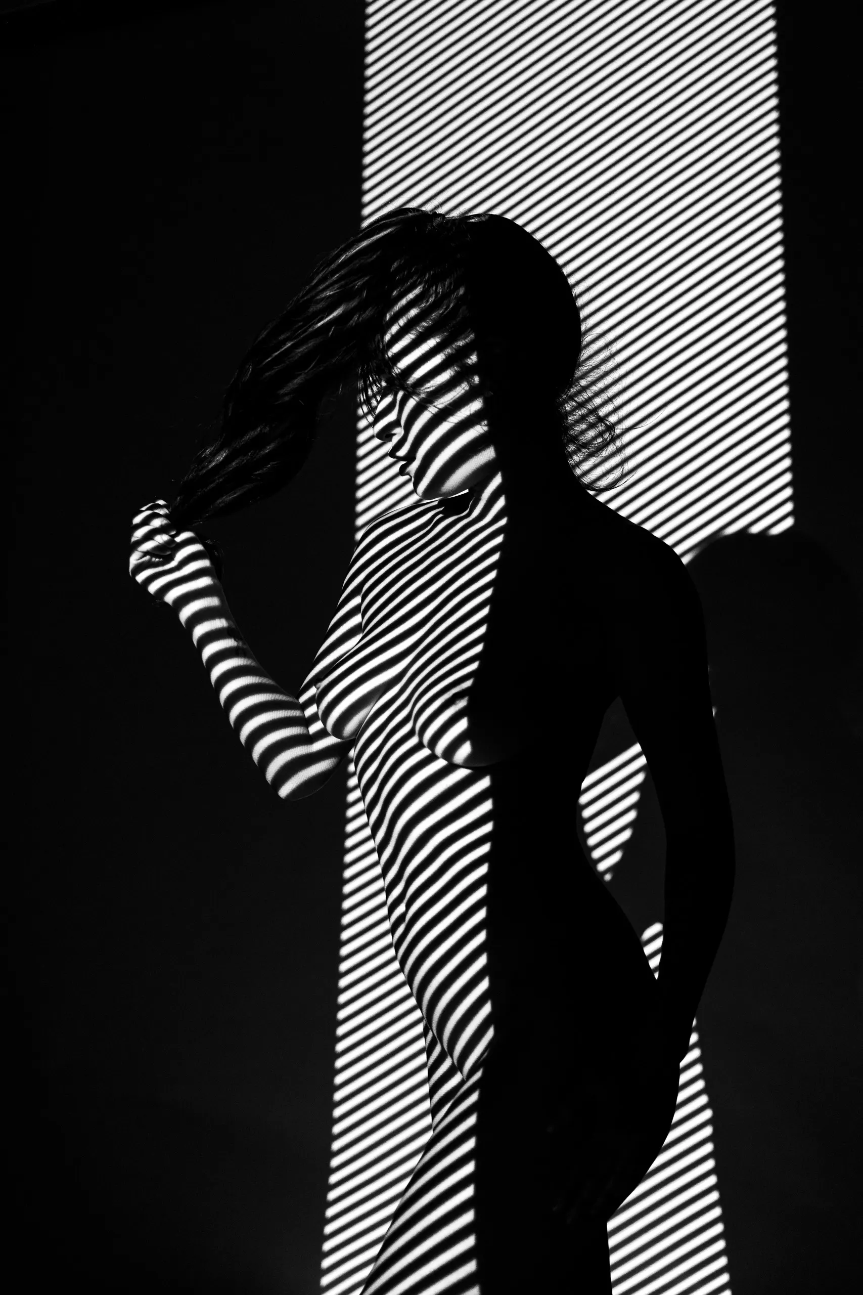 A woman stands in profile, pulling a hand through her hair, broad diagonal stripes of light and shadow wrapping her entire body against black.