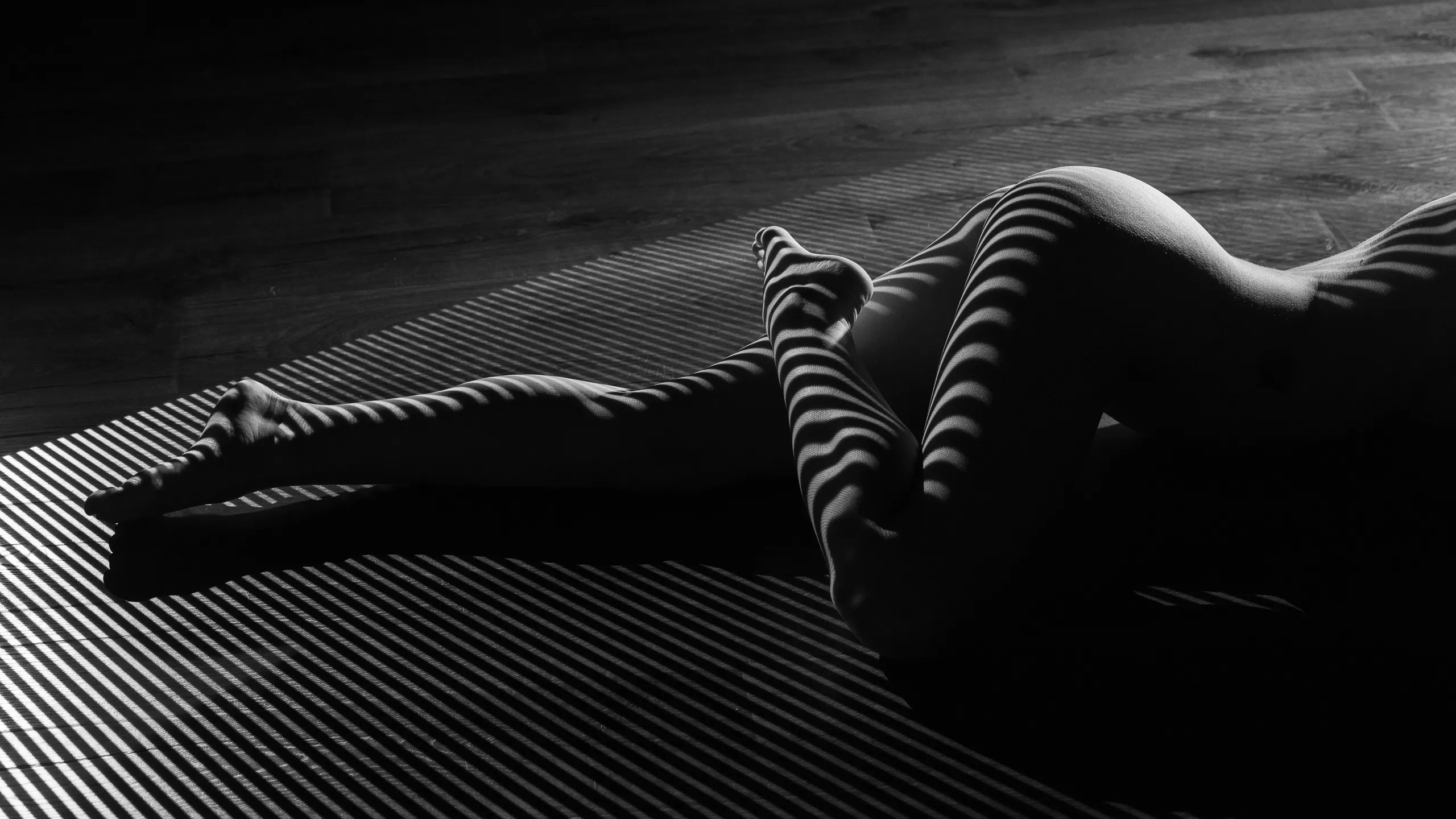 A close view of crossed legs and feet, striped light and shadow banding across the skin at a sharp diagonal against a dark wood floor.