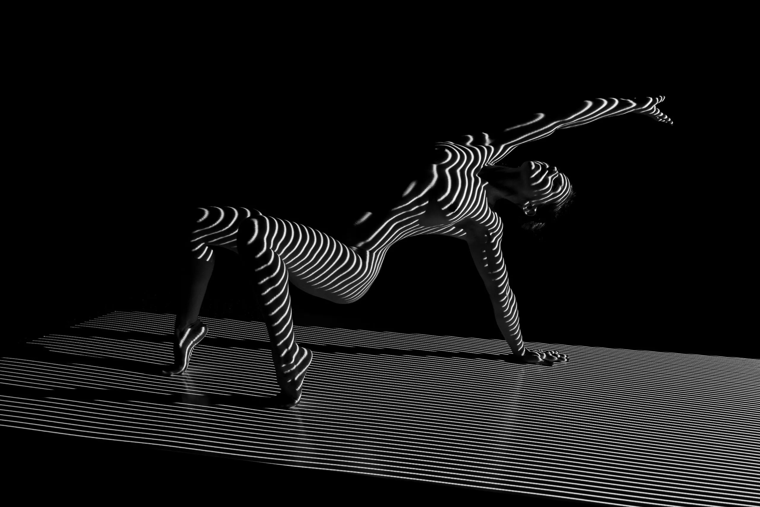 A woman posing on the floor, holding herself up by her legs and one arm, the other arm reaching behind her, black and white lines projected onto her skin and the floor.