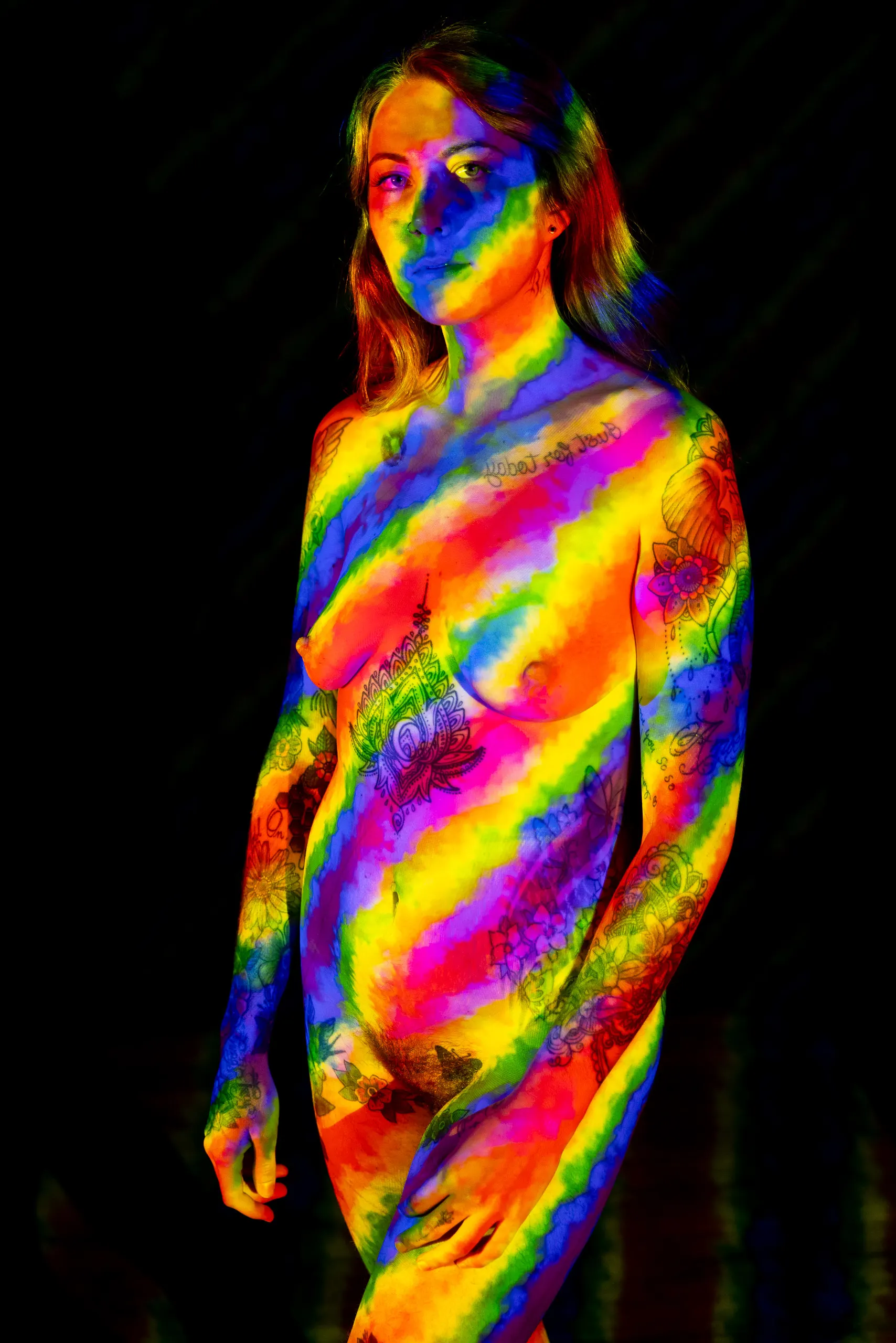 A woman stands with her arms at her sides, a vivid rainbow tie-dye pattern projected across her whole body, revealing an array of tattoos beneath the color.