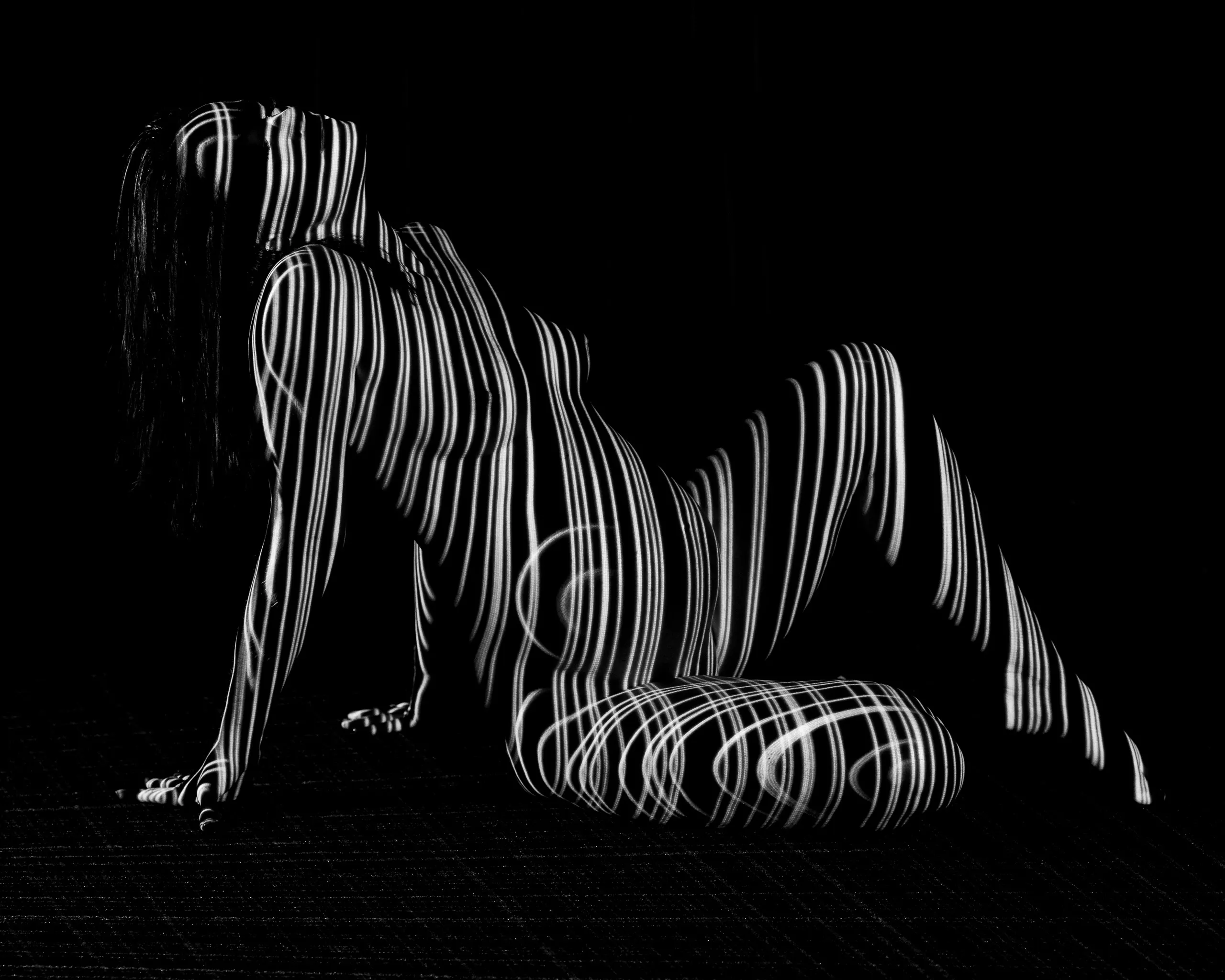 A pregnant woman sitting on a black floor, vertical stripes of projected light banding across her whole body against black.