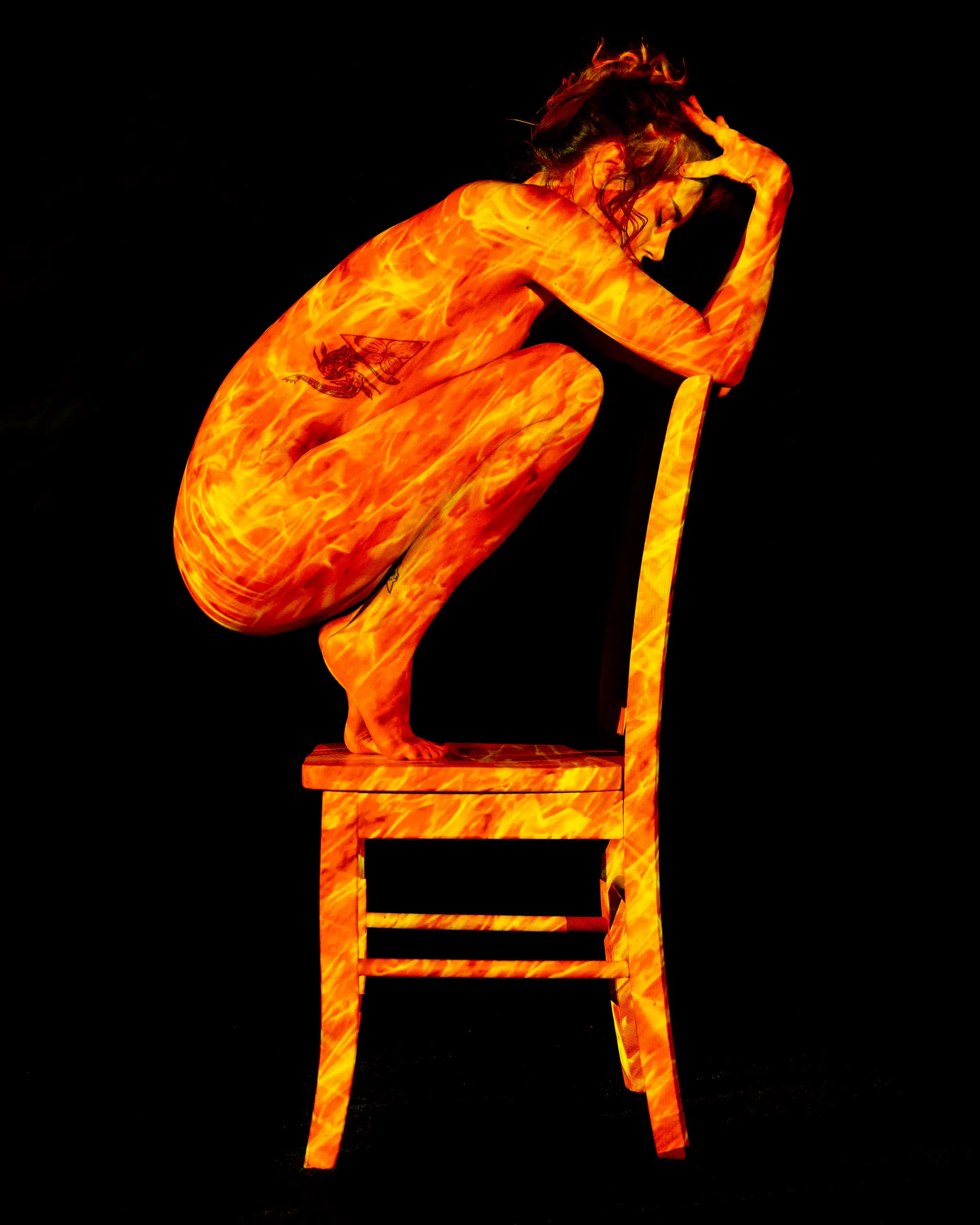 A woman crouches on a chair, curled tightly with her head resting on one arm, a rippling pattern of fire-orange light covering her whole body and the chair beneath her.