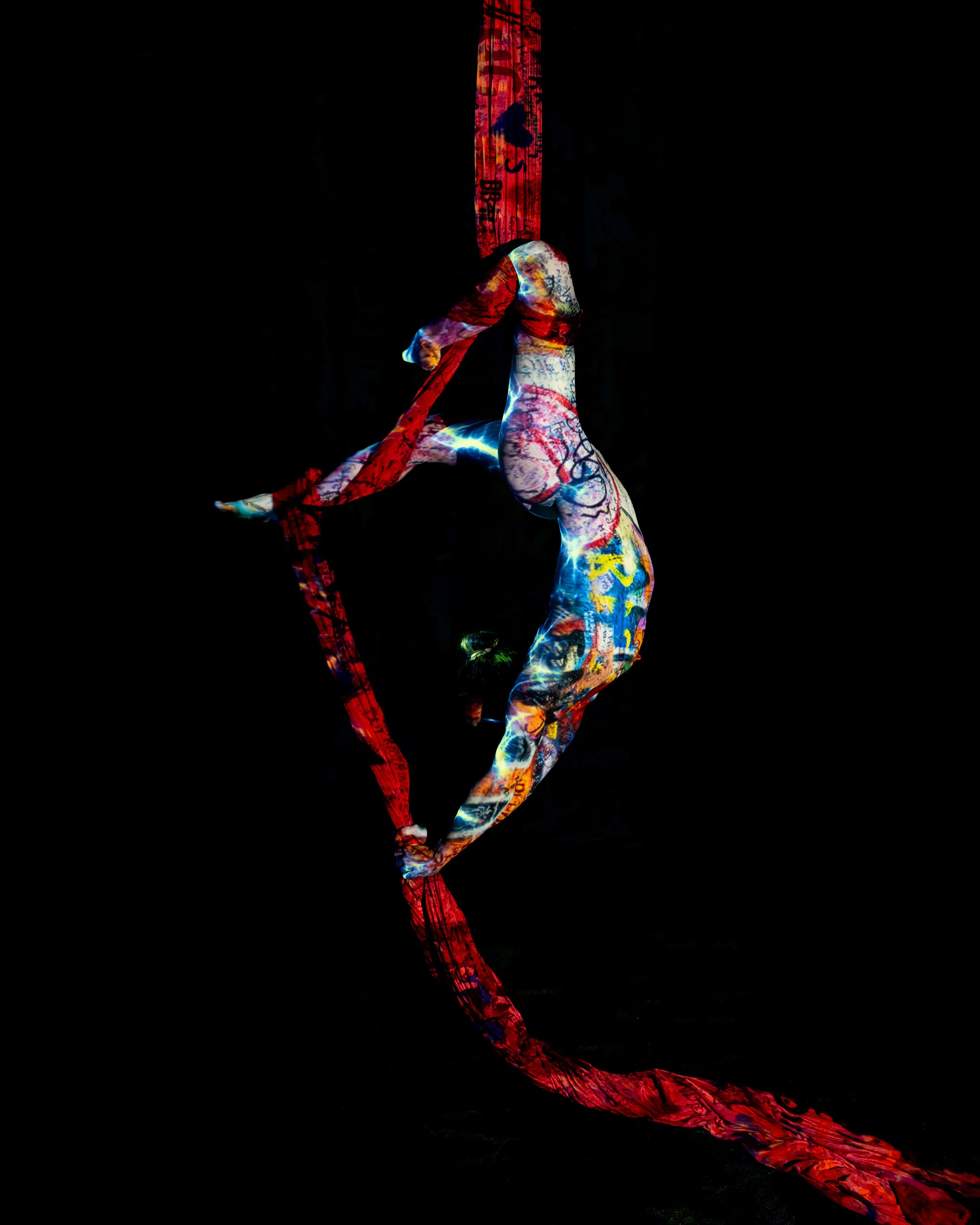 A woman hangs from red aerial silks, one leg extended, a colorful graffiti-style pattern of light and text projected across her body and the fabric alike.