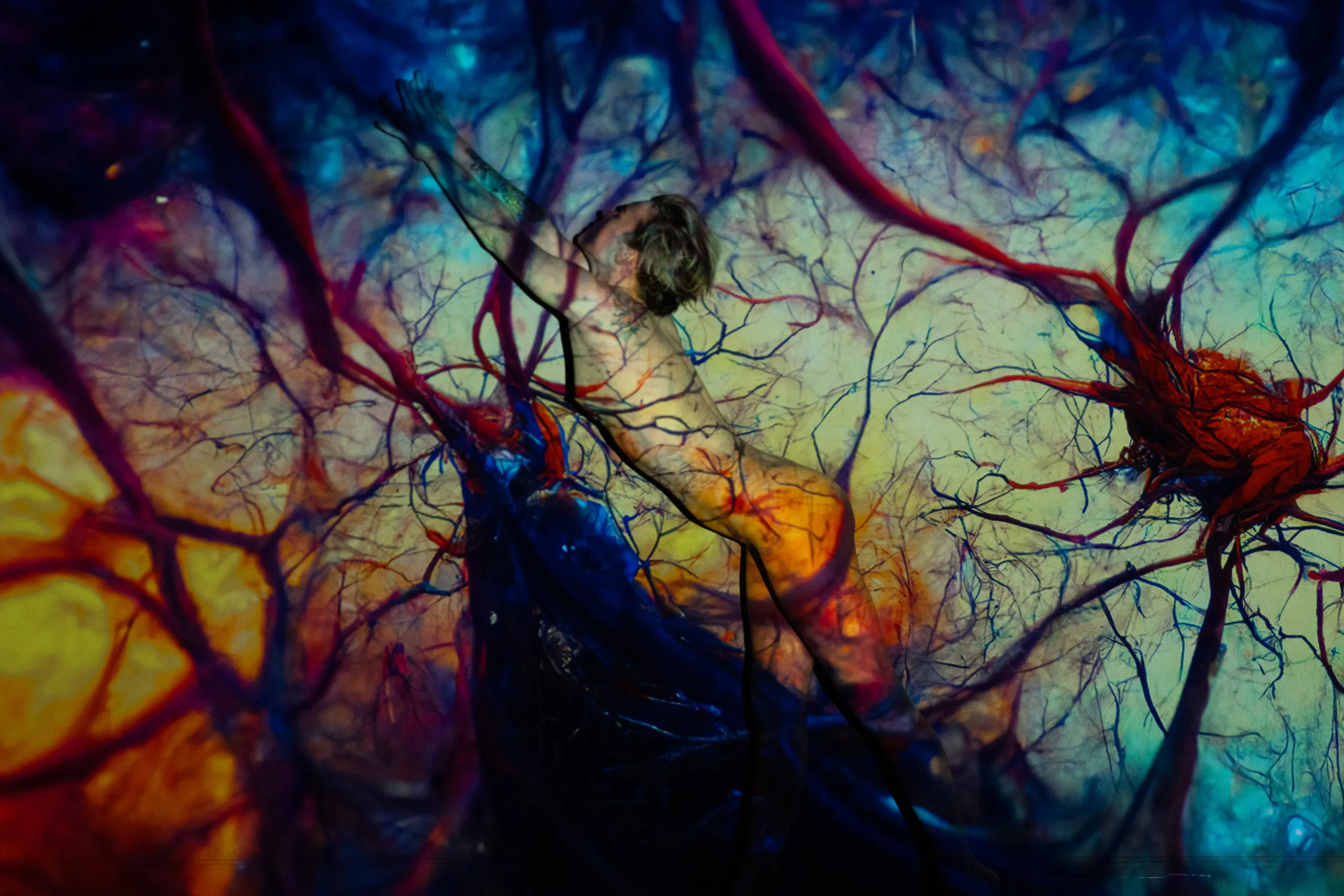 A woman leans forward with one arm reaching upward, a dense web of red and blue veinlike light projected across her body and the surrounding space.