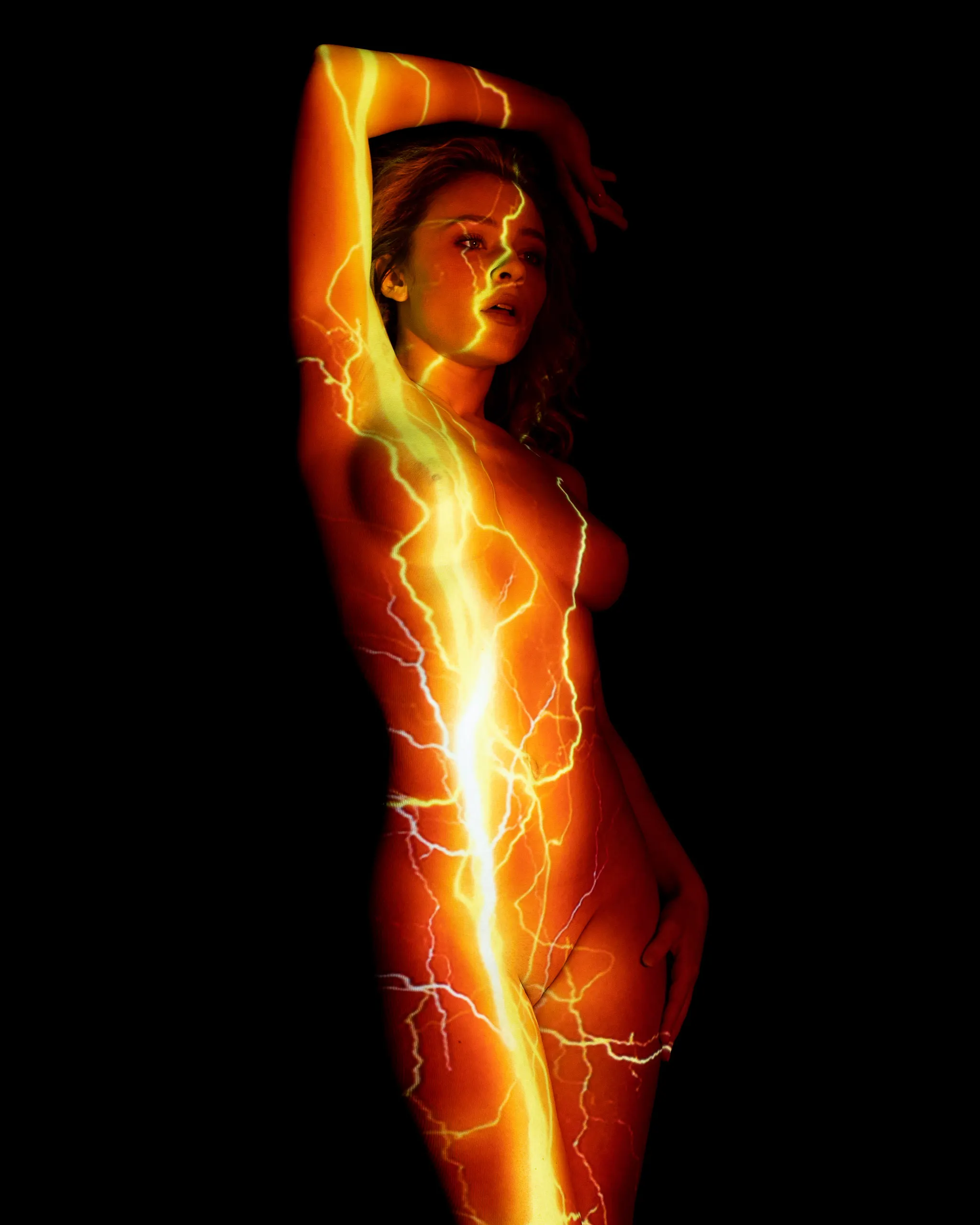 A woman stands with one arm raised overhead, jagged bolts of golden lightning projected down the length of her body against black.