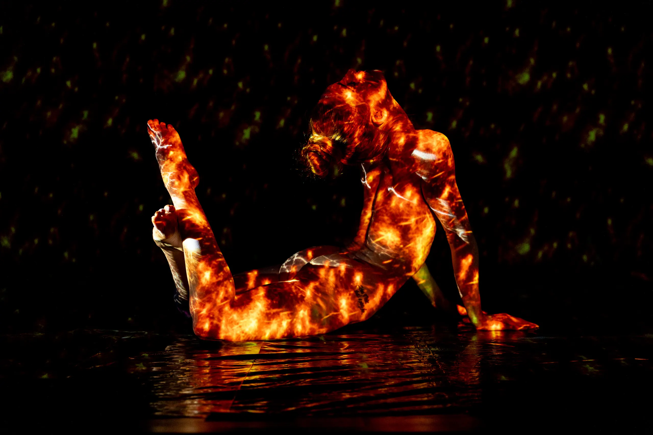 A woman arches backward with her legs raised, a fiery, crackling pattern of orange and gold light covering her entire body against black.