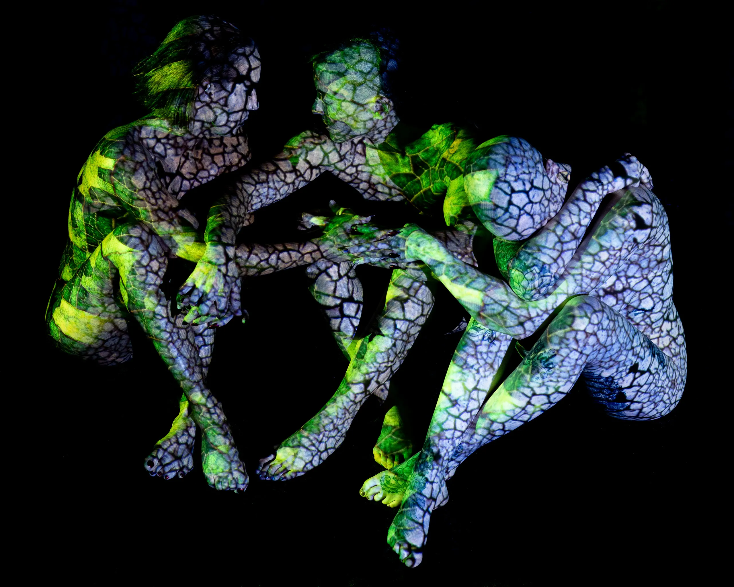 Three women seated on the floor, a mottled cracked pattern of light in green, yellow, and white covering their bodies against black.