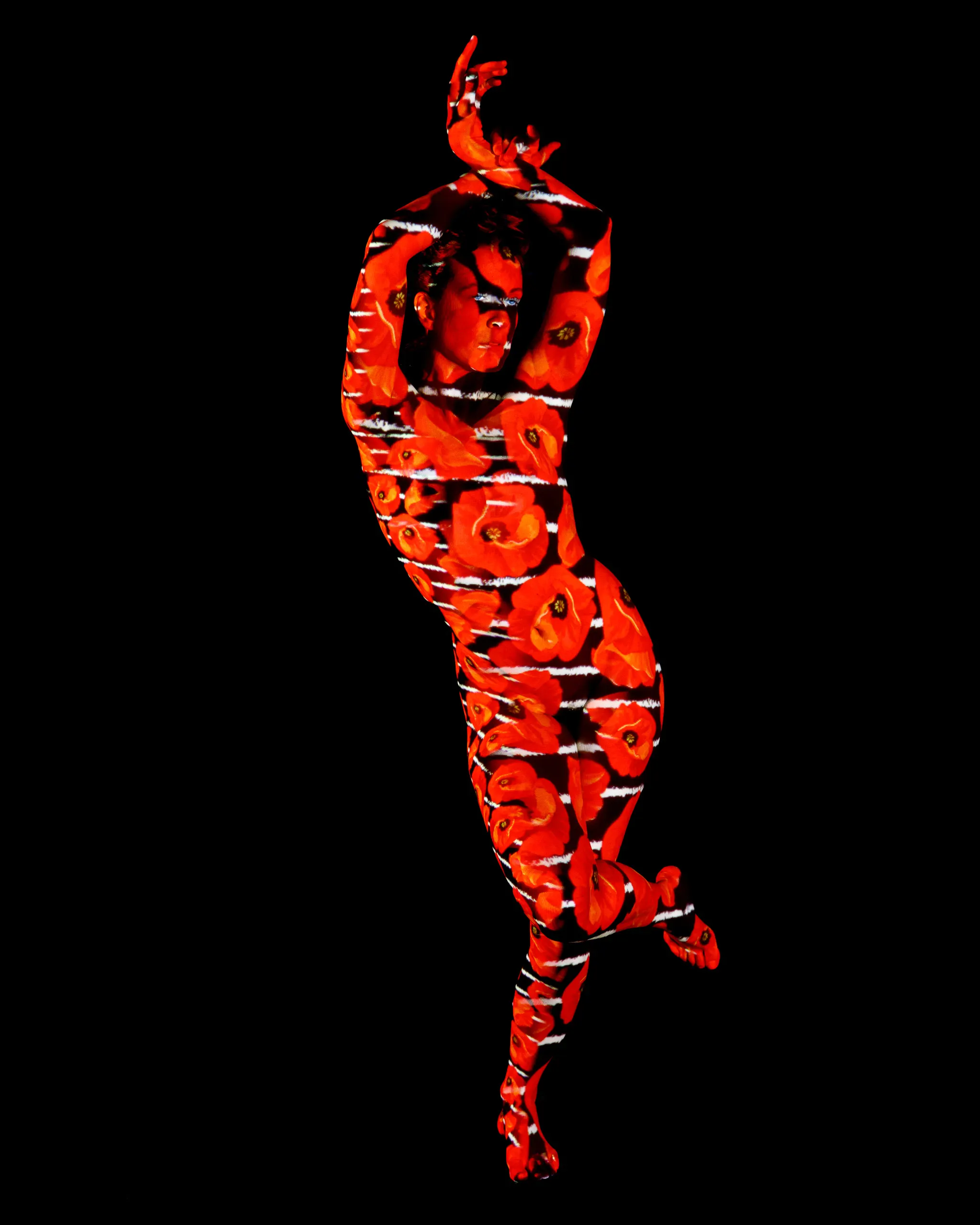 A woman poses with both arms raised overhead, oversized red poppy blossoms projected across her whole body in horizontal bands against black.
