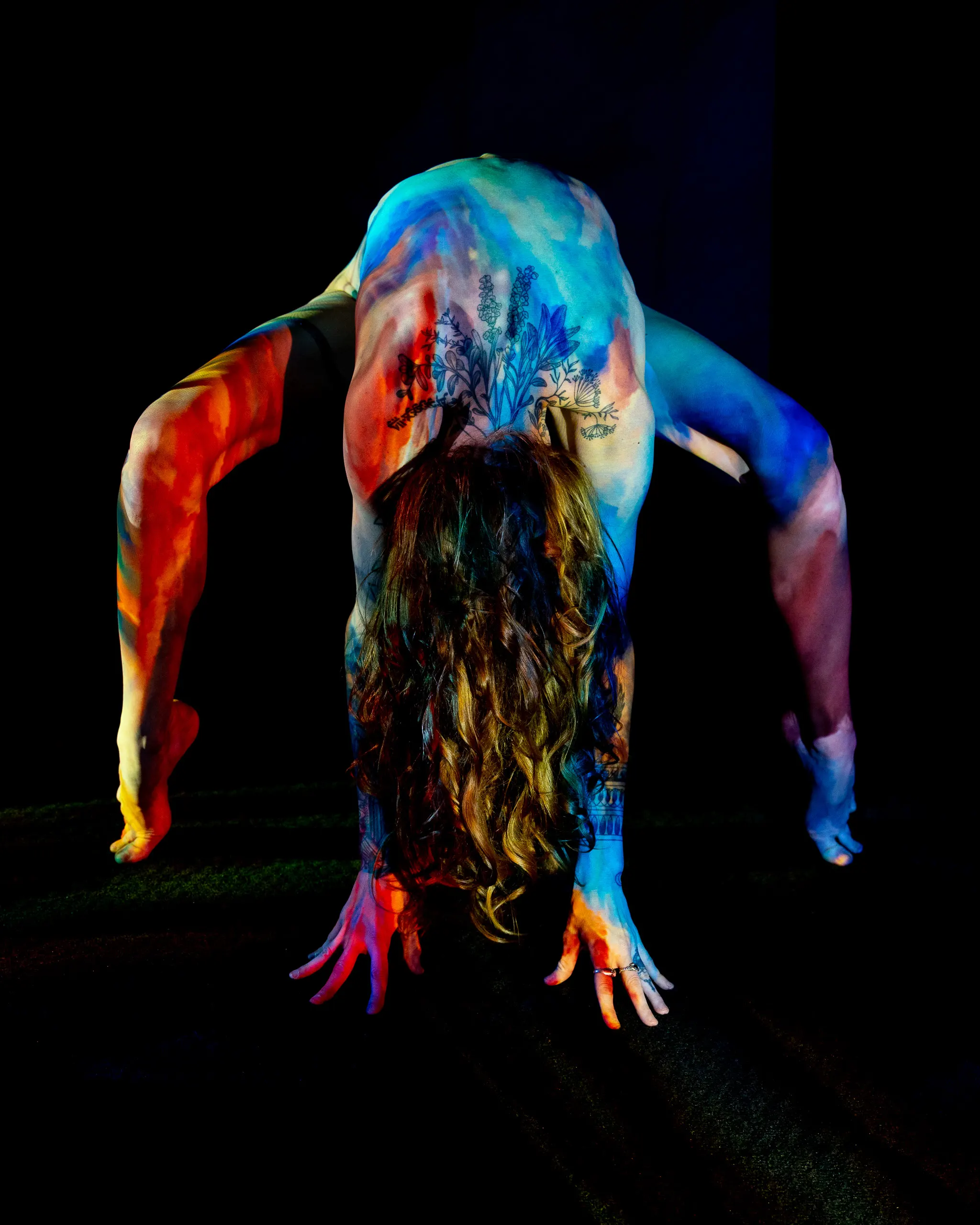A woman bends forward with her hair falling loose, warm orange and cool blue watercolor-like light washing across her back and arms, floral tattoos visible on her back.