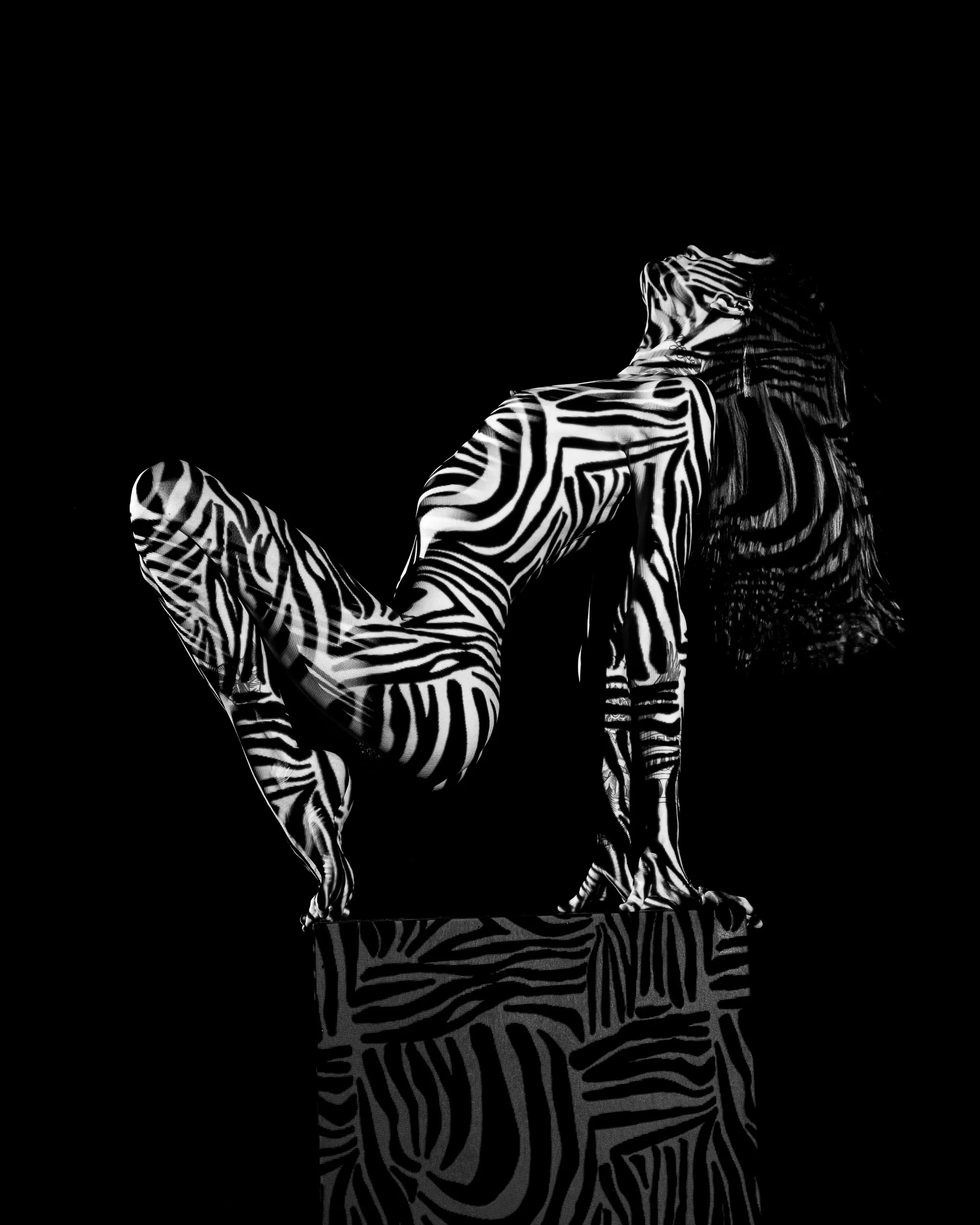 A woman kneels on a pedestal, arching back with one leg raised, bold zebra-stripe patterns of light covering her whole body and hair against black.