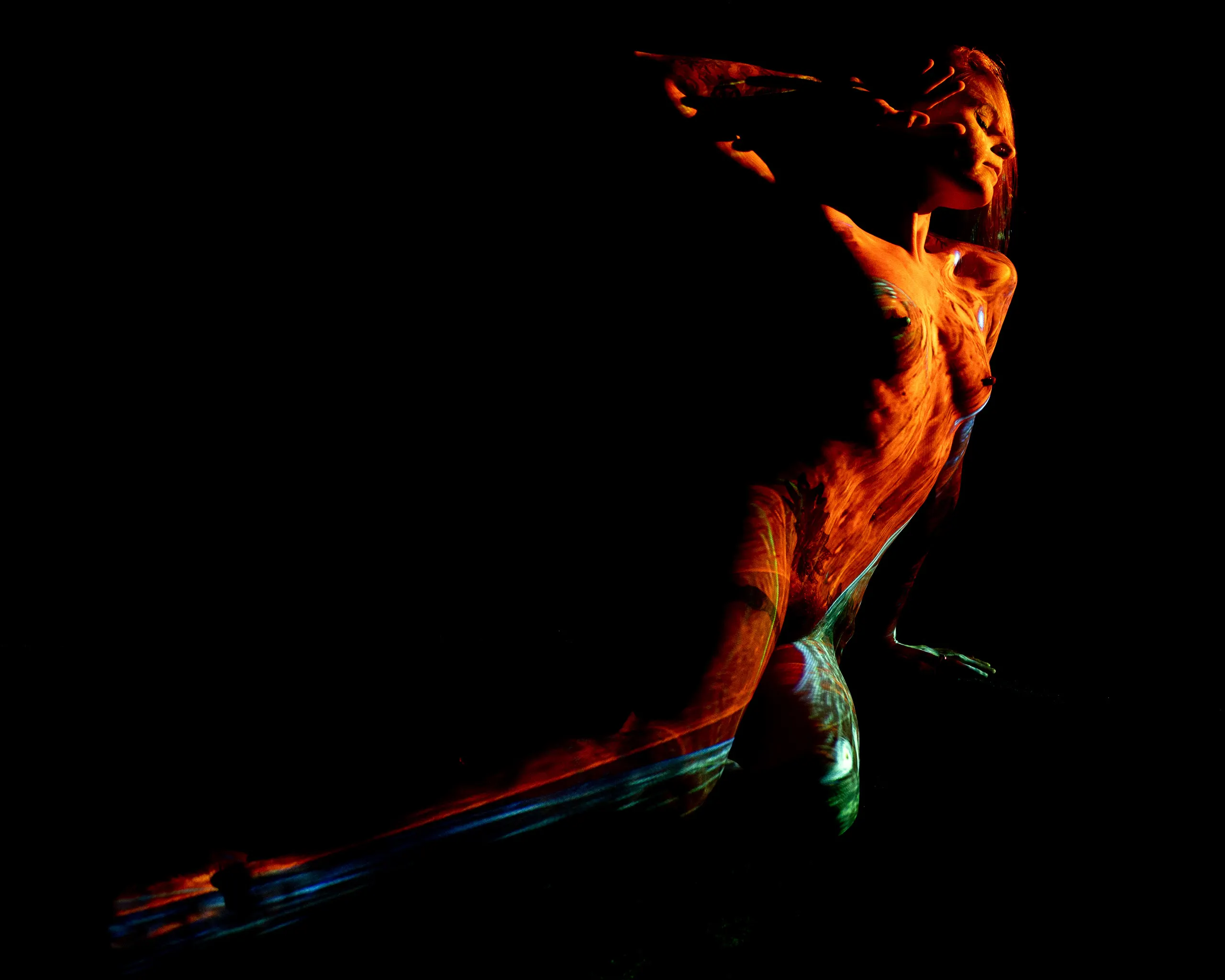A woman leans back with one arm raised overhead, warm orange and cool teal projected light washing across her body against black.