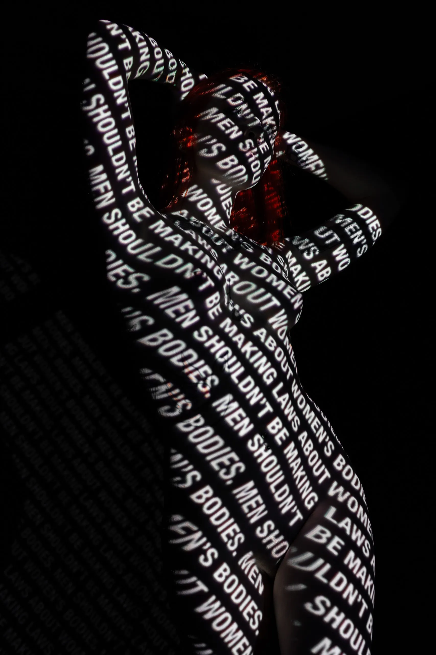 A woman stands with both arms raised, bold text projected across her entire body reading statements about bodily autonomy, set against black.