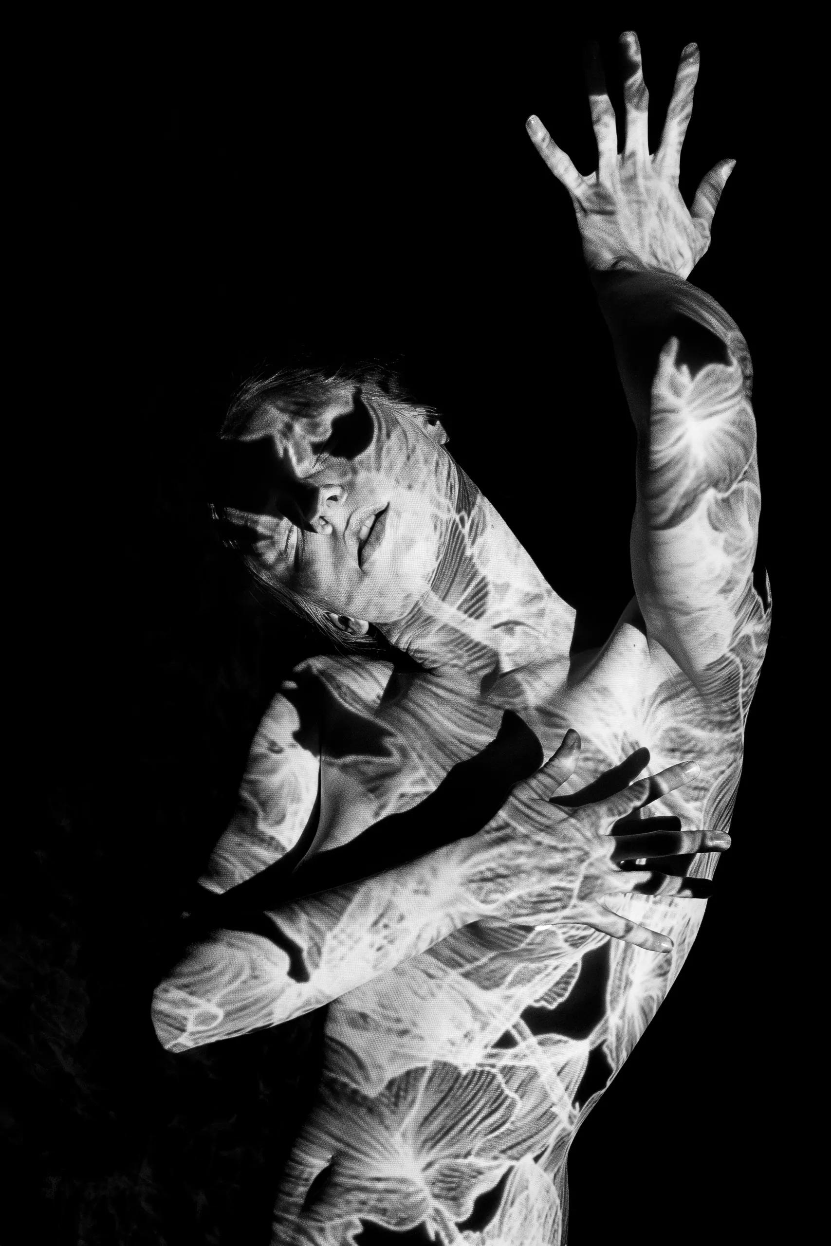 A woman reaches one hand upward, the other pressed to her chest, delicate white floral and vein-like patterns of light covering her body against black.