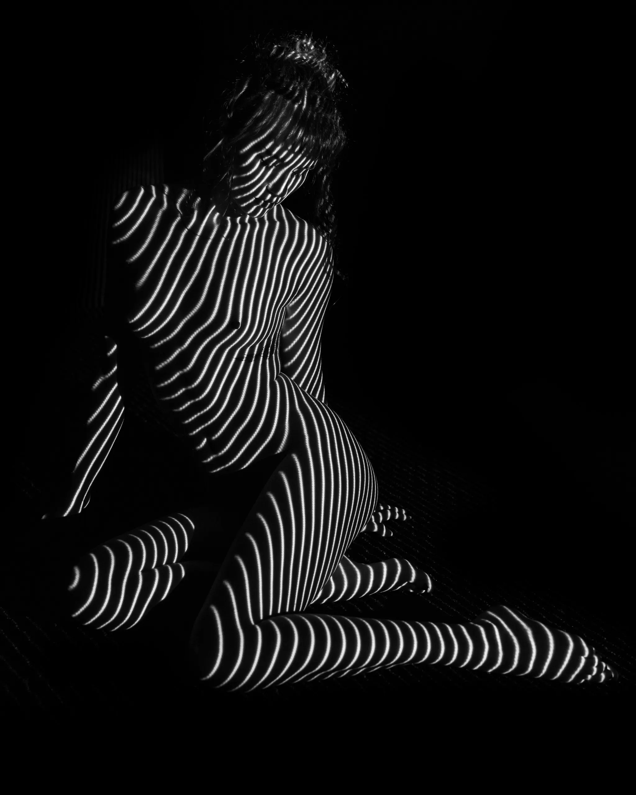 A woman sits on the floor with her legs kicked out, curving lines of light wrapping around her torso, hip, and legs against black.