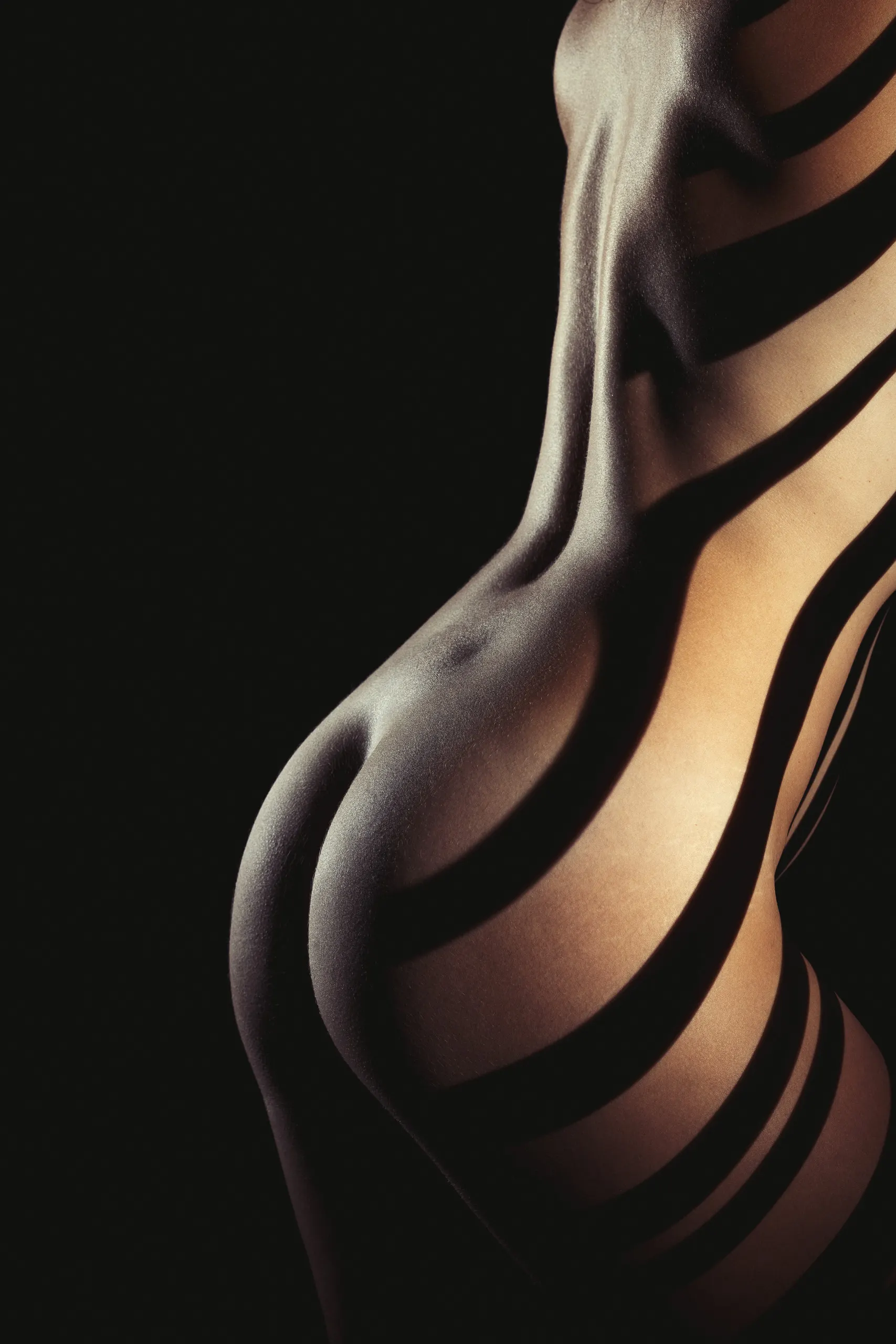 A close, low-light study of a hip and torso in profile, broad bands of shadow wrapping the body's curve like ribs of light. Warm skin tones show through the gaps.
