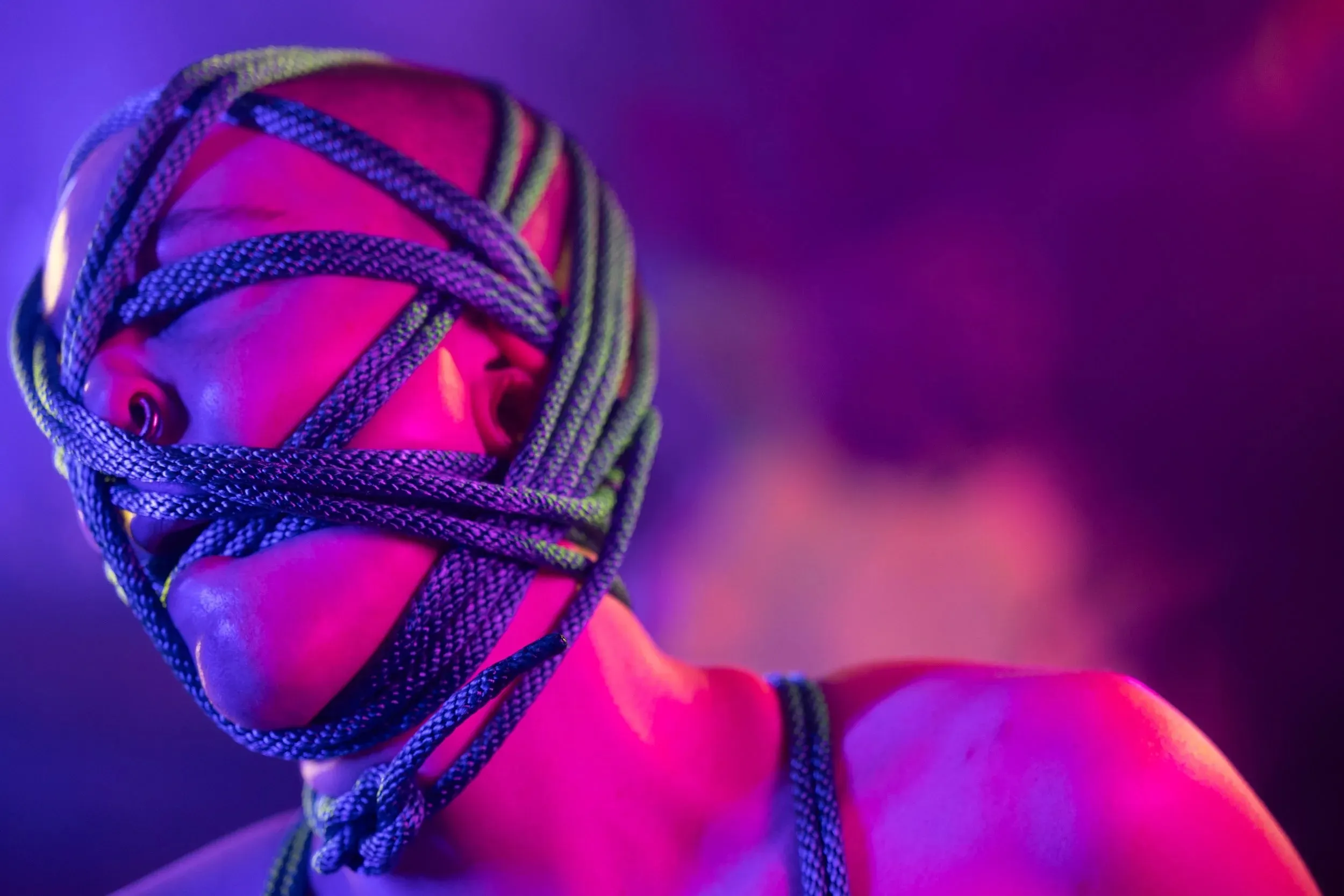 A rope-covered face turns slightly in saturated purple light.