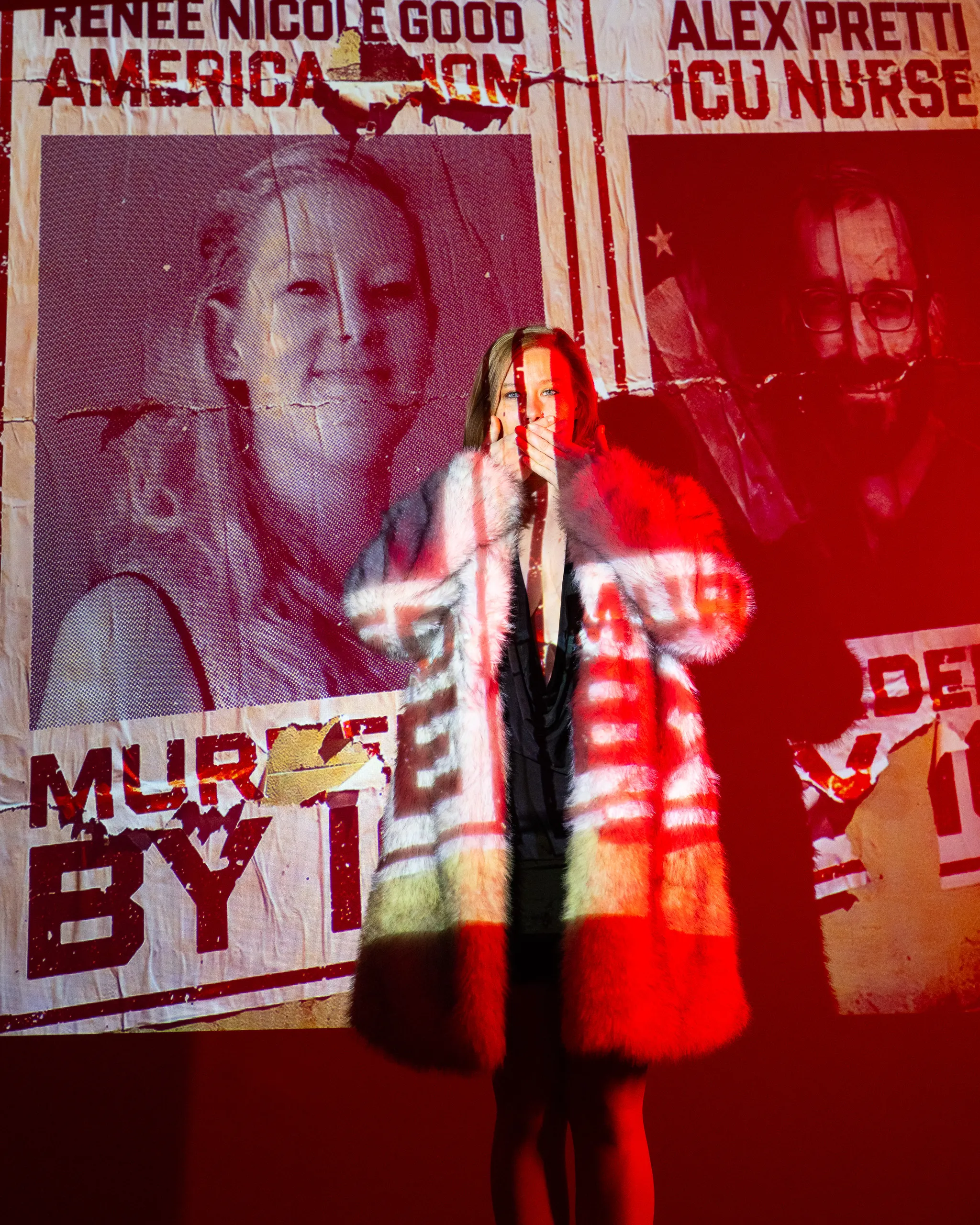 A woman stands in red light with portraits and news headlines projected onto her body and the wall.