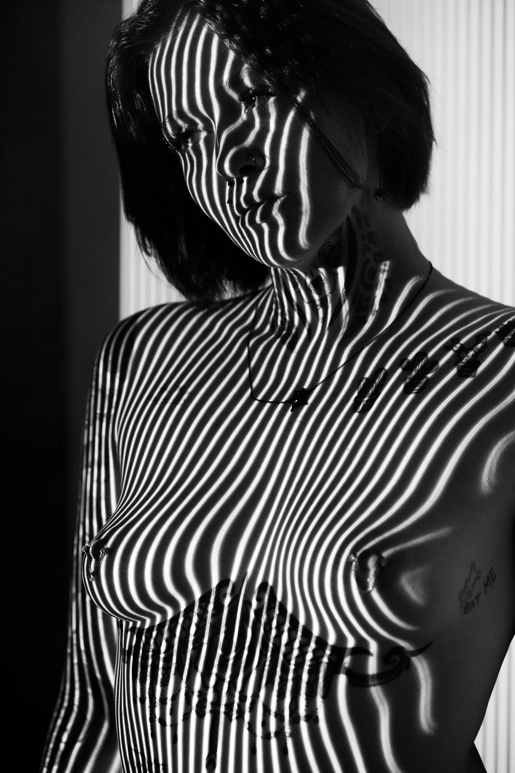 A woman tilts her head beside a white v-flat as thin vertical stripes of light wrap her face and torso, black and white.