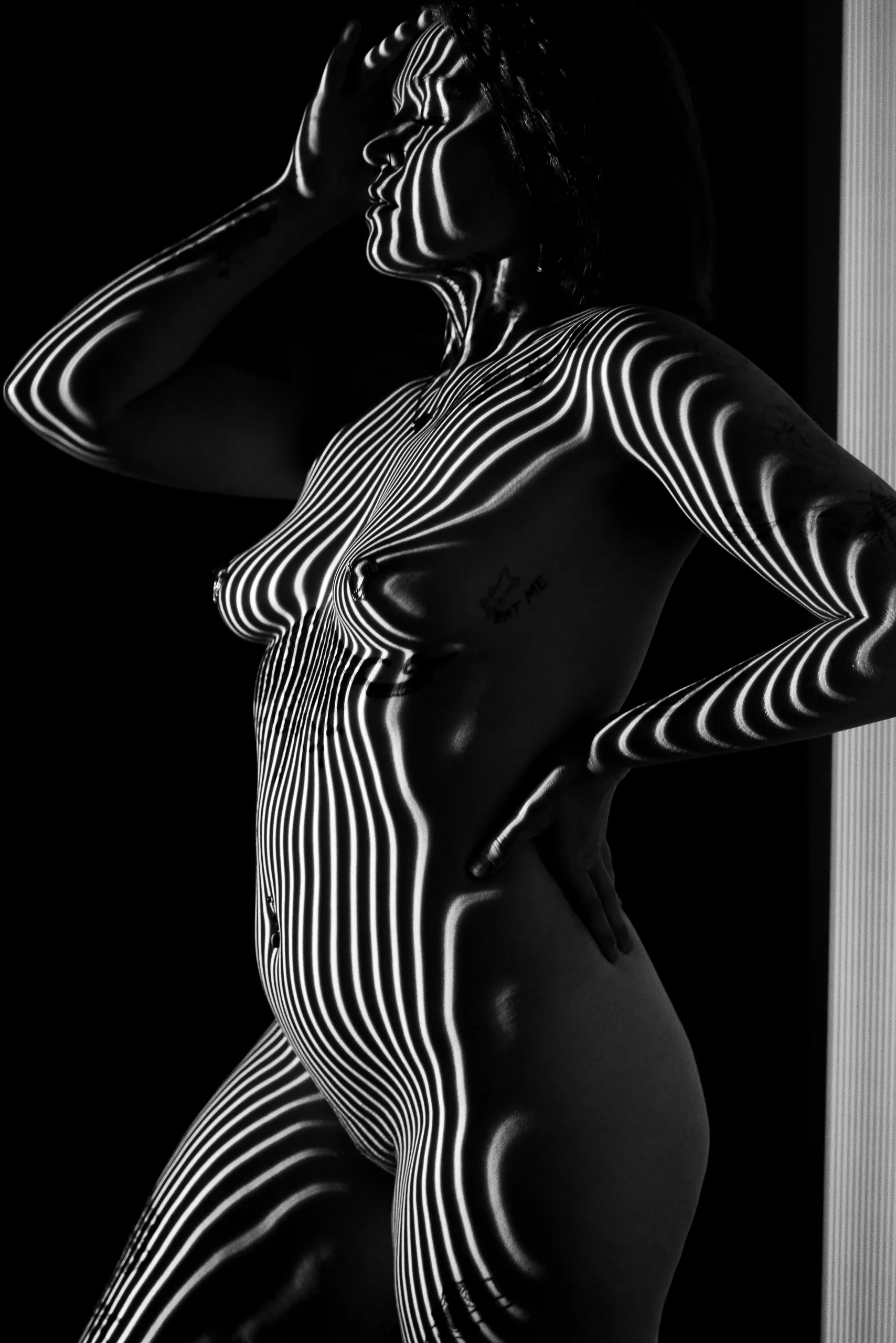 A standing woman in profile raises one hand to her head, her body traced by curving projected stripes against a black background.