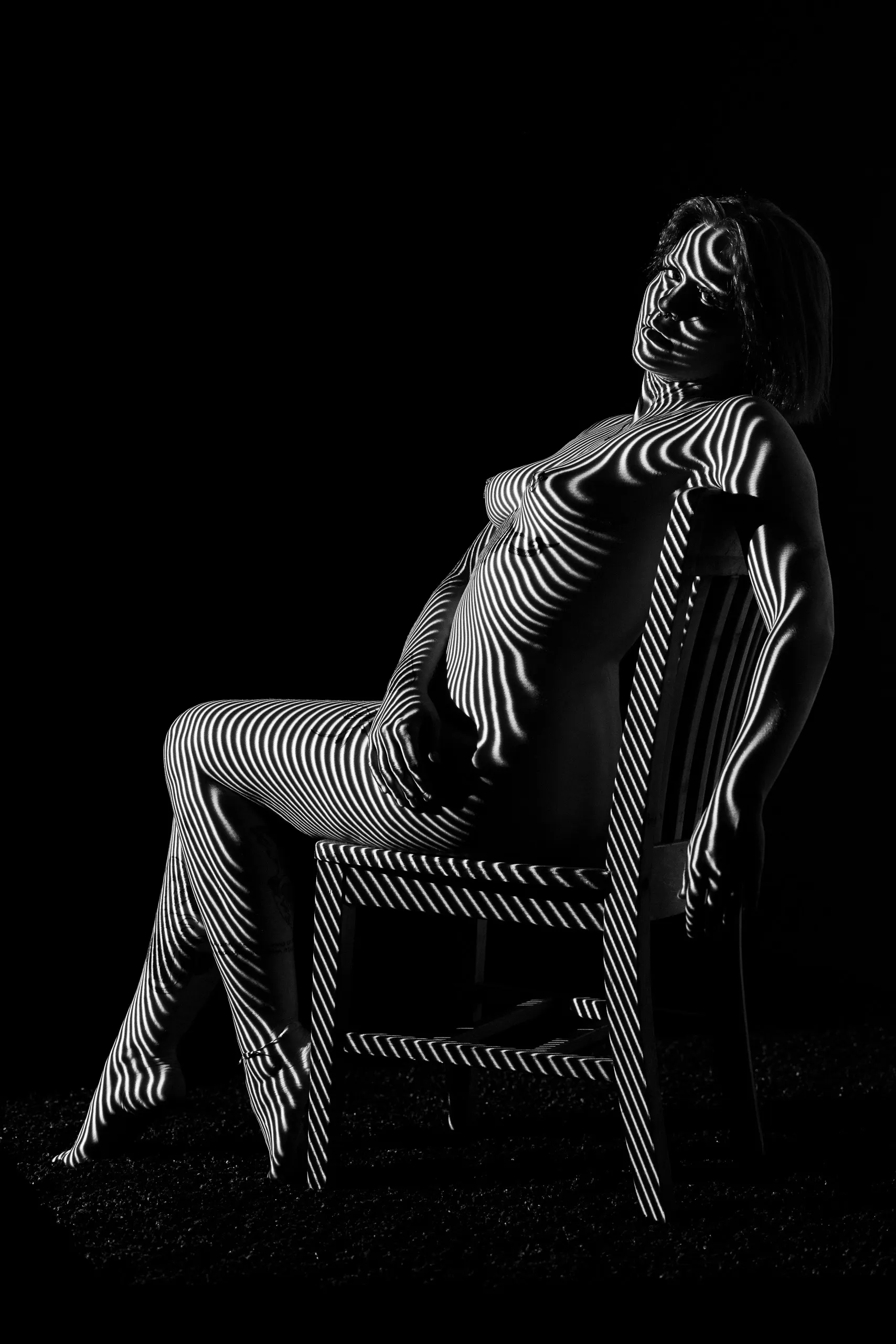 A woman leans back in a wooden chair in a dark room, projected lines bending across her body and the chair.