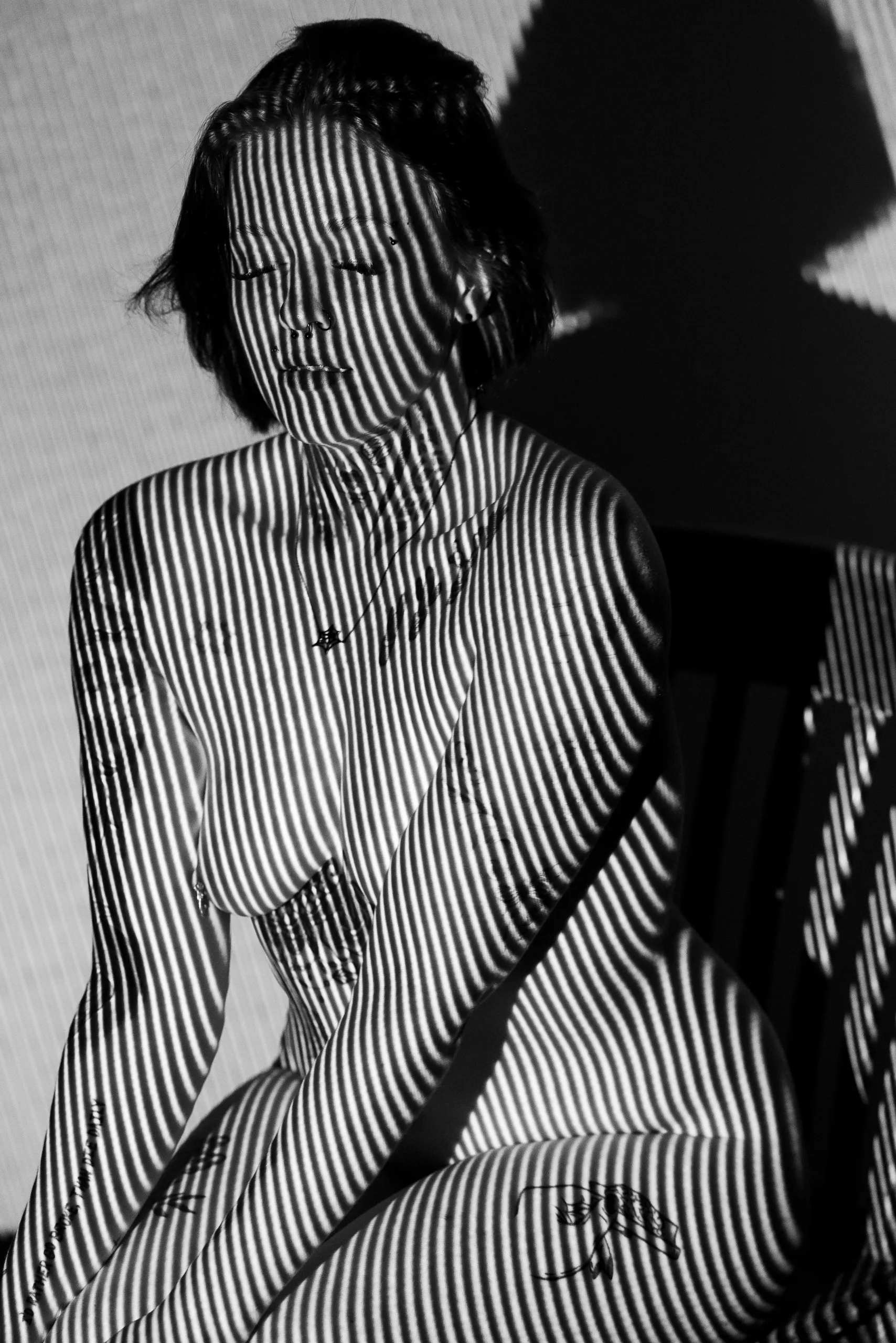 A seated woman with closed eyes is covered in thin vertical stripes, her shadow cast on the white v-flat behind her.