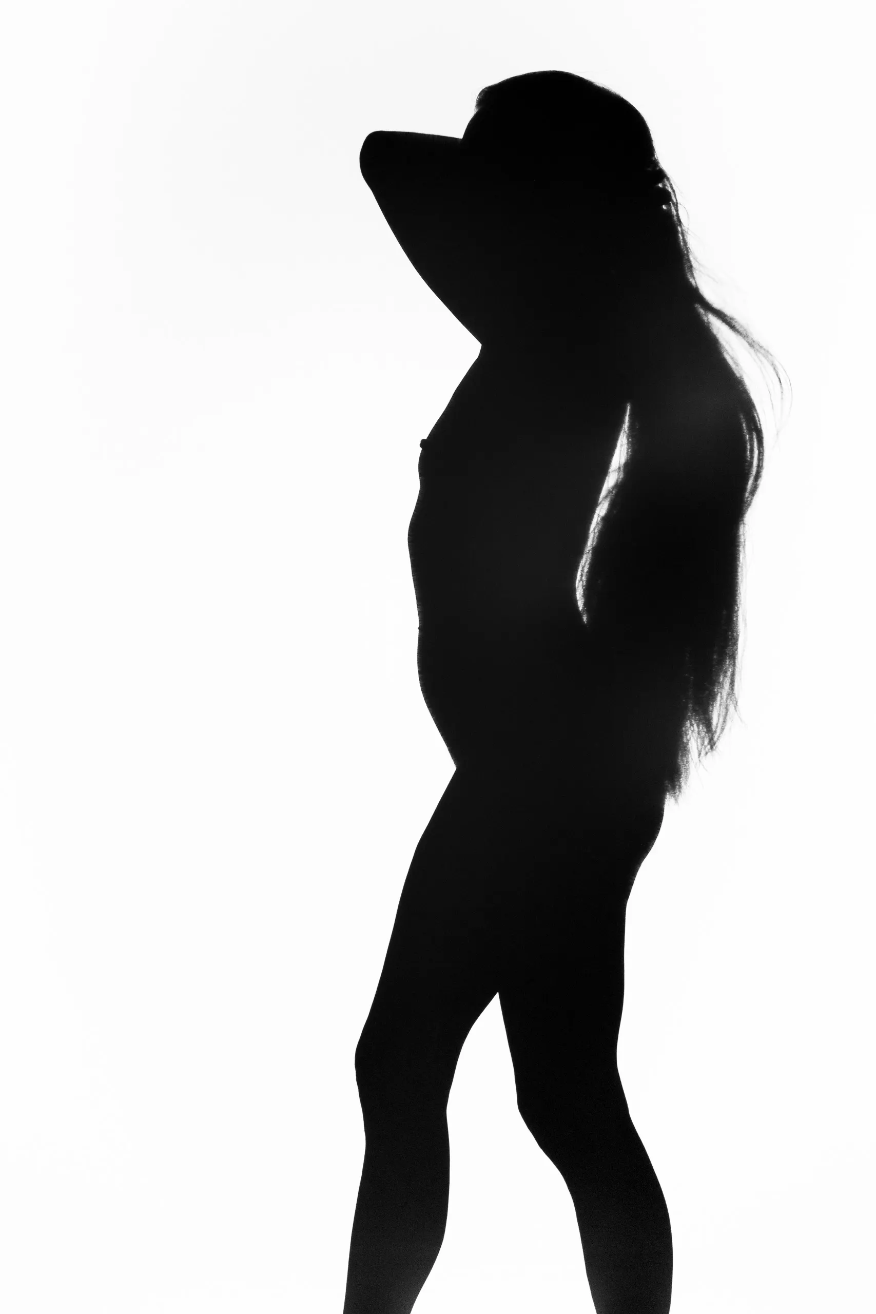 A silhouetted woman turns partly away with one arm bent behind her head, a bright rim of light tracing her back and the long hair spilling over her shoulder.